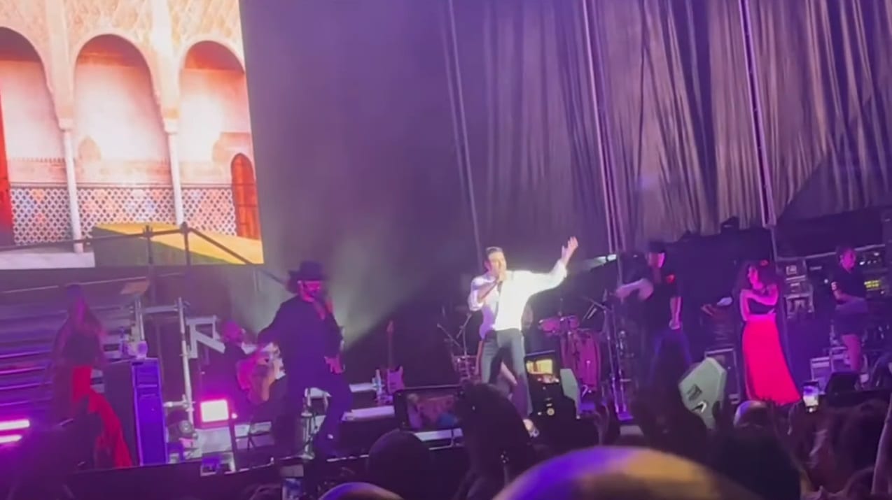 Un cantante con camisa clara levanta un brazo y canta frente a un micrófono, con una banda detrás bajo luces moradas.