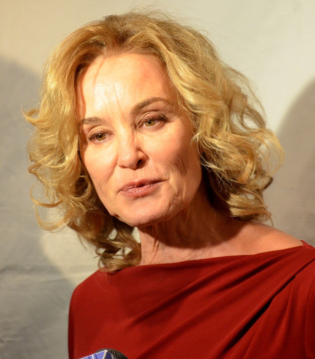 Jessica Lange sonríe frente a fotógrafos en una alfombra roja, con vestido oscuro y el cabello suelto.