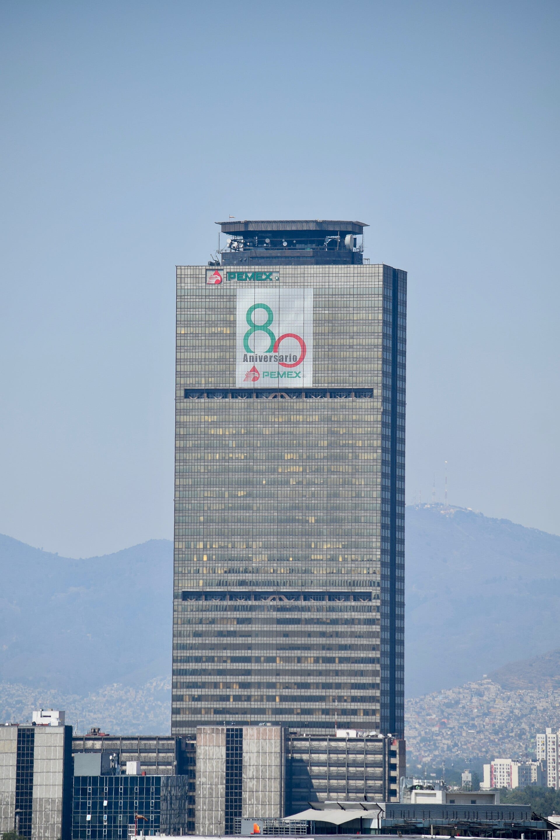 La Torre Ejecutiva de Pemex, con el logotipo de la empresa y un cartel de aniversario, vista desde abajo con montañas al fondo.
