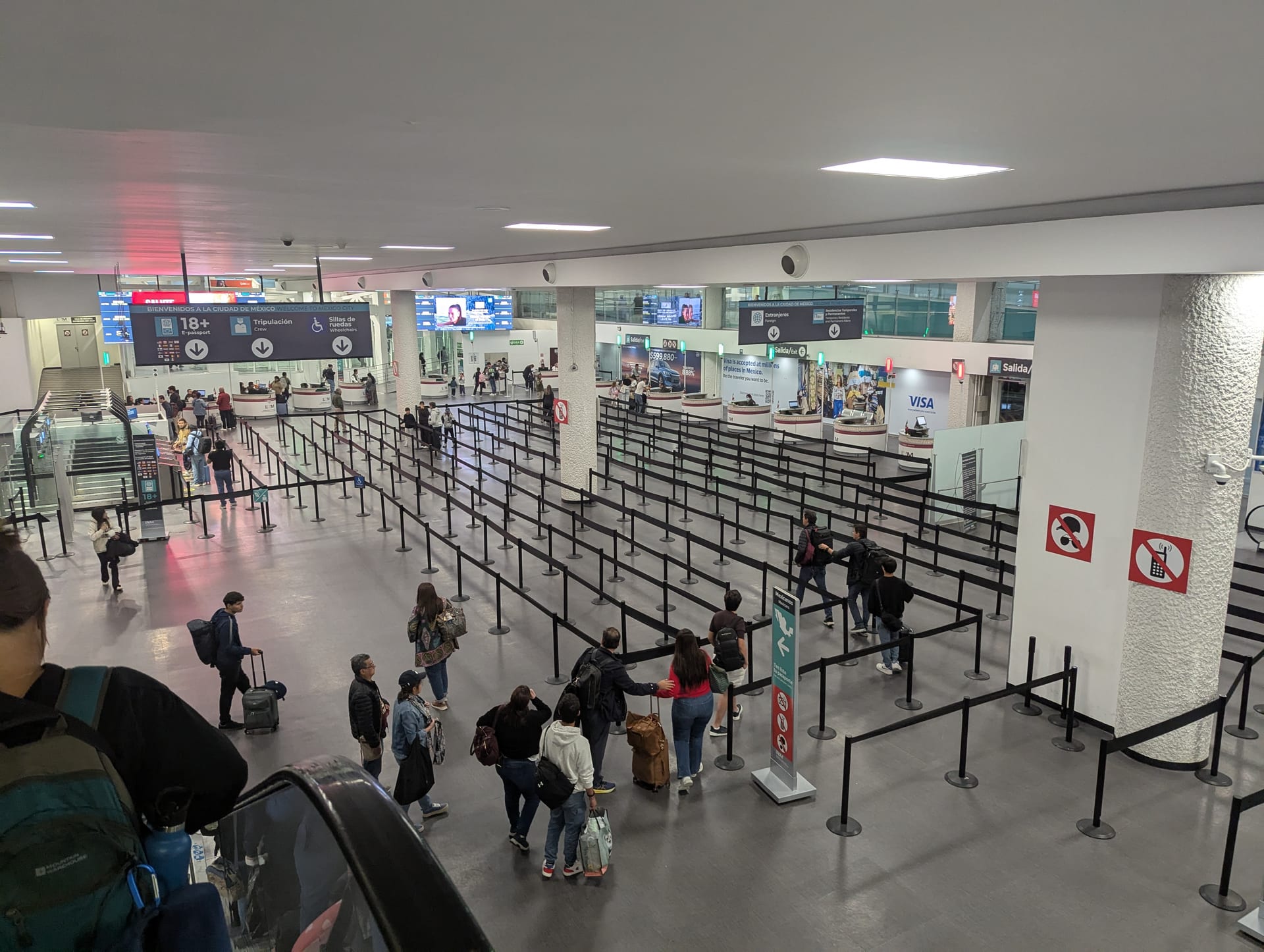 Viajeros hacen fila entre postes con cinta guía en el área de llegadas de una terminal aérea, bajo pantallas de señalización en español e inglés.