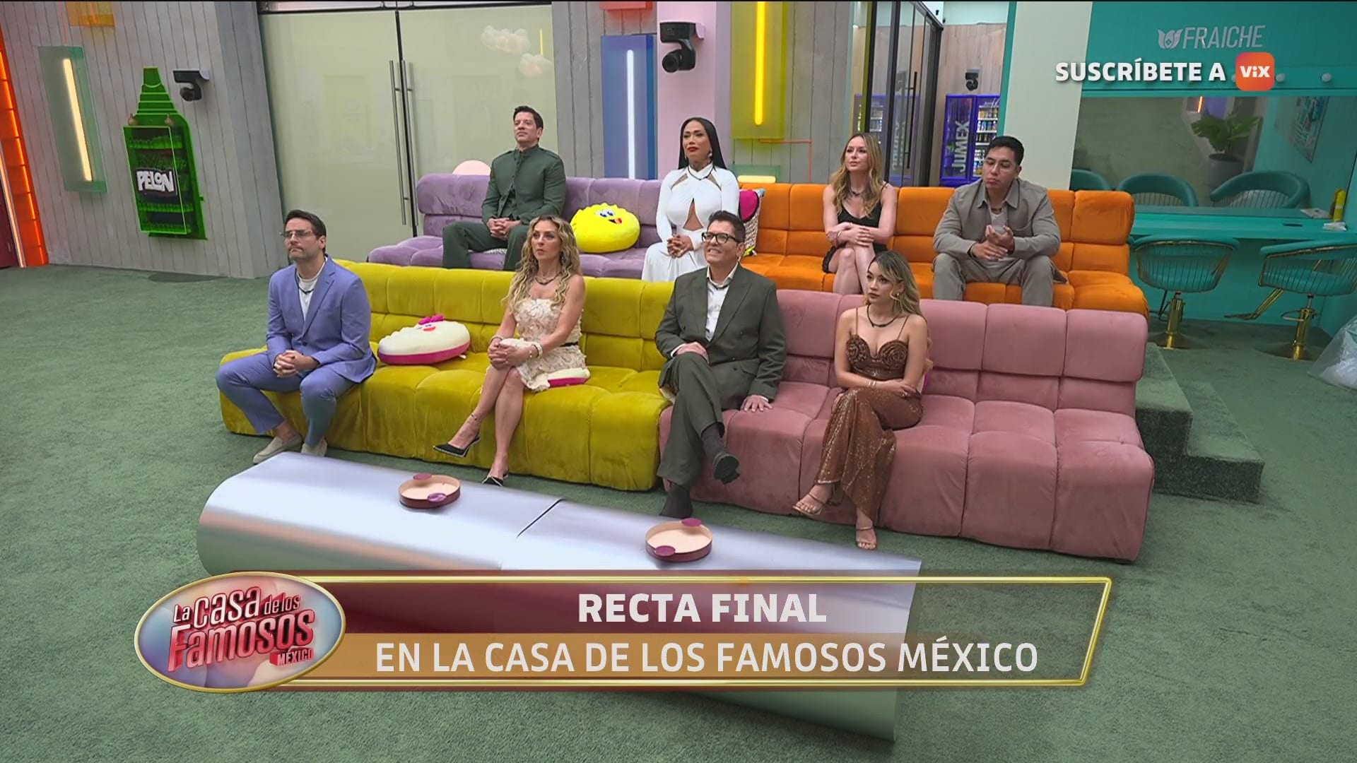 Ocho habitantes vestidos de gala están sentados en sillones de colores dentro de la casa durante una transmisión en vivo.