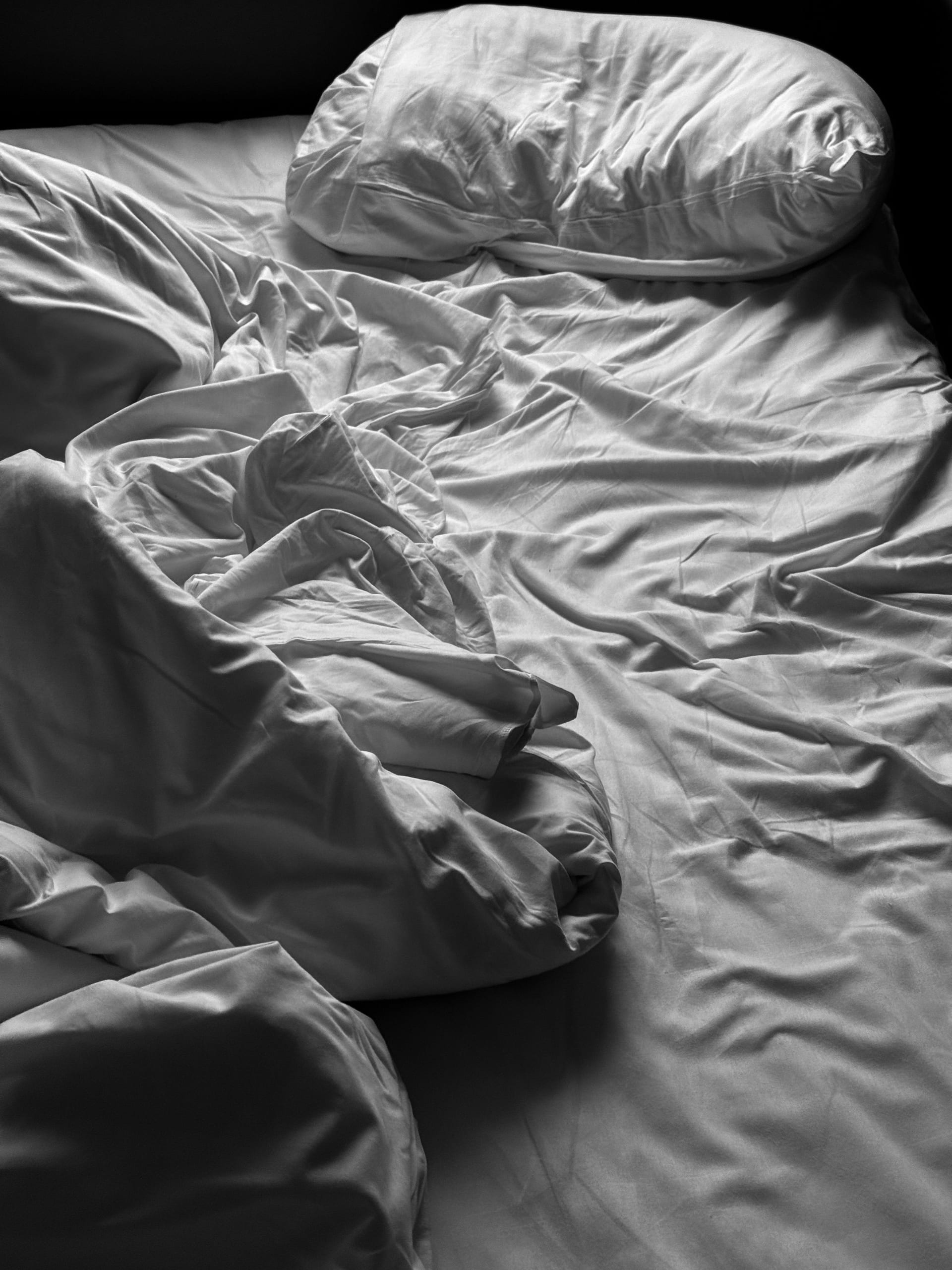 Cama deshecha con sábanas y almohadas blancas arrugadas, fotografiada en blanco y negro con luz lateral.