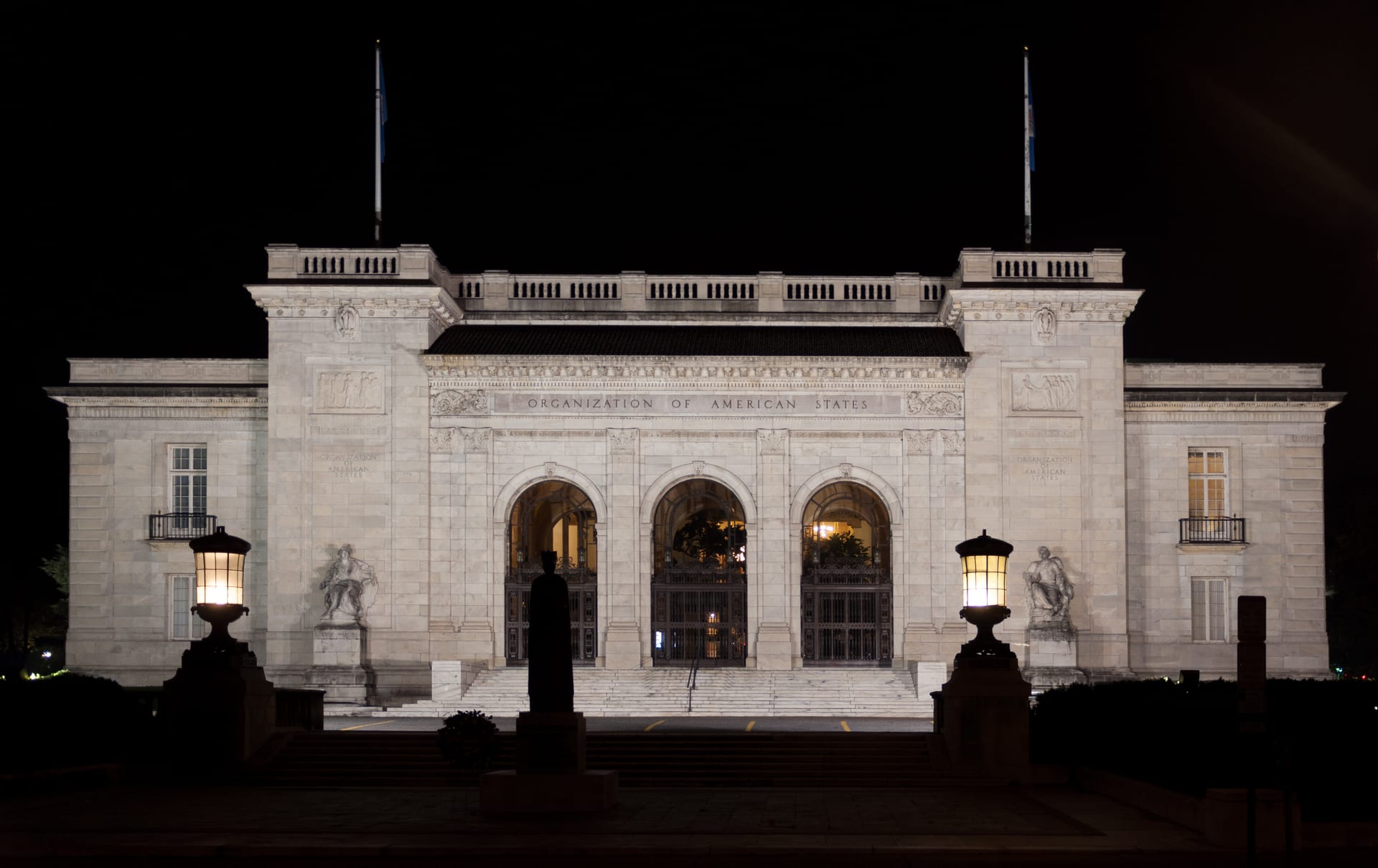 Fachada nocturna de un edificio con tres arcos de entrada, columnas y el letrero Organization of American States, flanqueada por estatuas y faroles encendidos.