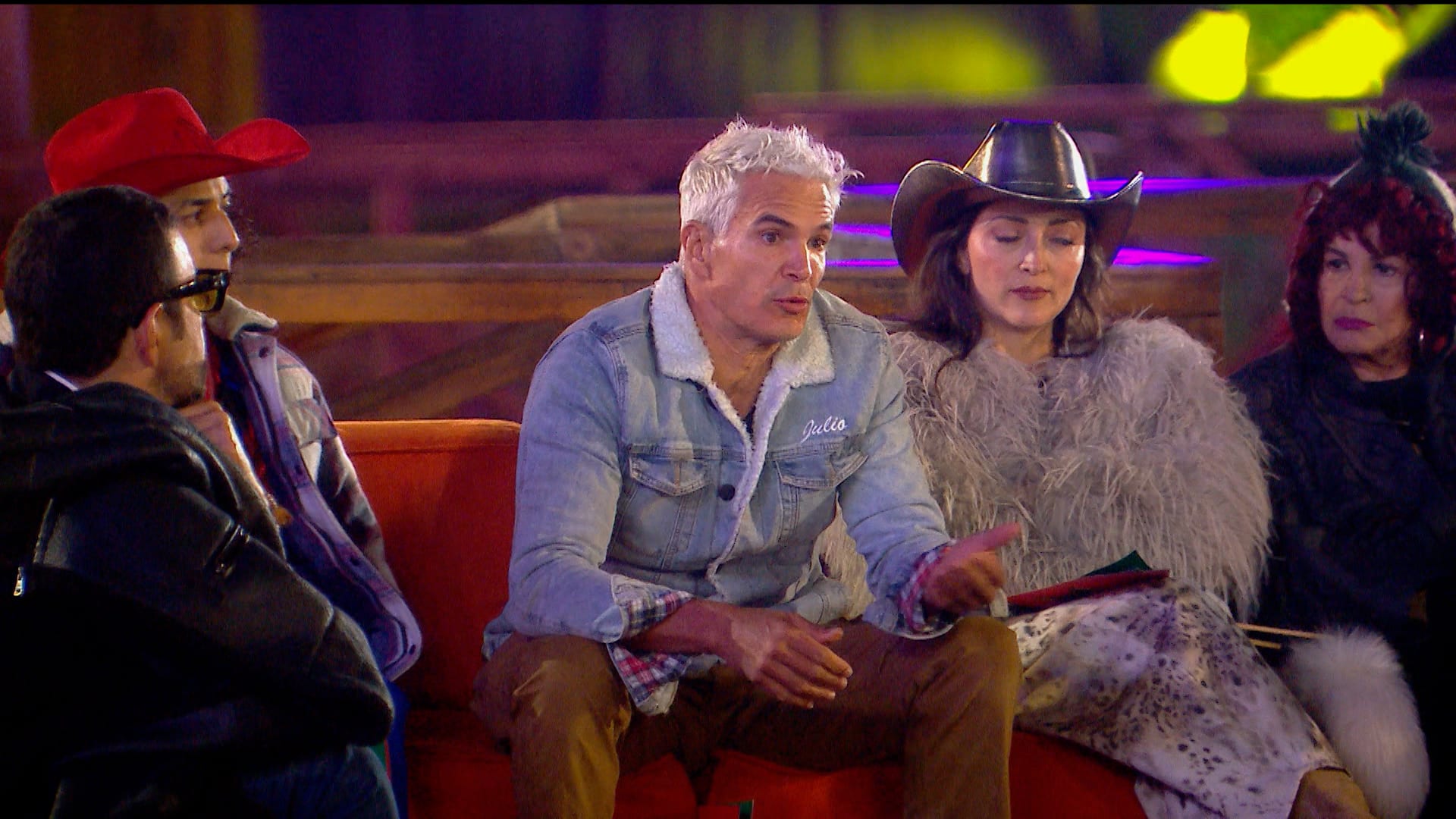 Cuatro integrantes del reality conversan de noche en sillones, uno con sombrero rojo y otro con sombrero vaquero negro.