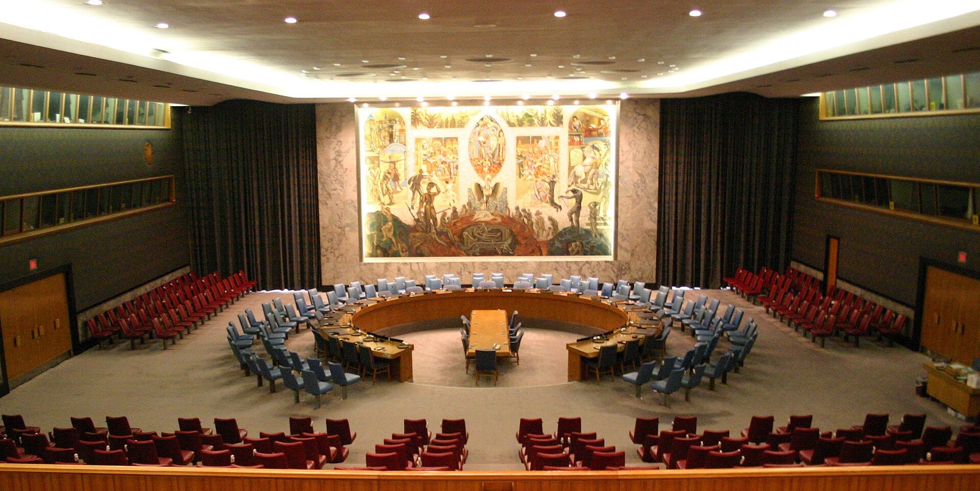 Sala circular con un mural de fondo y una mesa en herradura rodeada de sillas vacías.