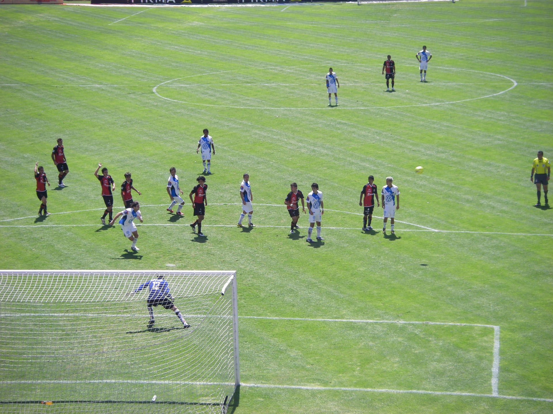 Futbolistas con uniforme blanco y azul, y otros de rayas rojas y negras, disputan un balón cerca del área durante un partido de Liga MX.