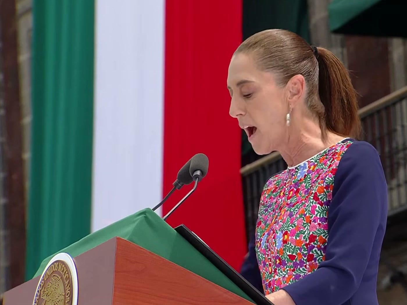 Claudia Sheinbaum lee su discurso en un atril, con la bandera de México y un edificio de cantera al fondo.