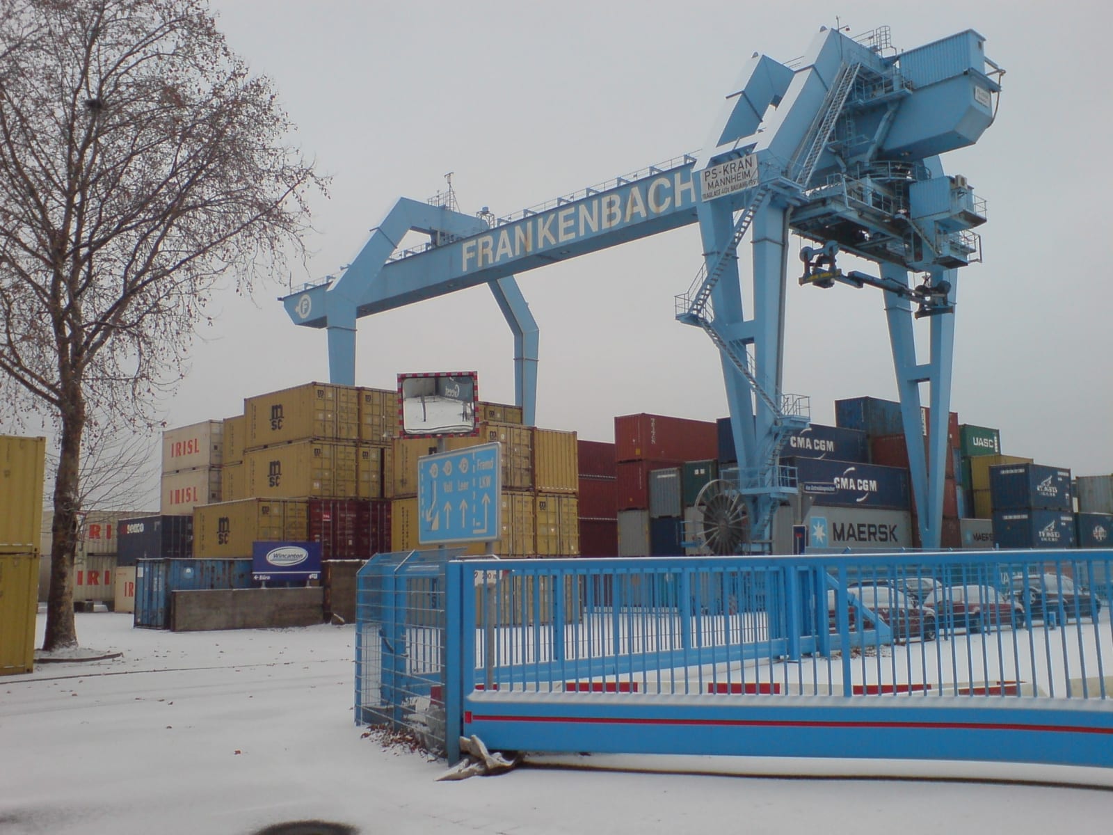 Una grúa pórtico azul levanta contenedores de carga apilados en una terminal portuaria con nieve en el suelo.
