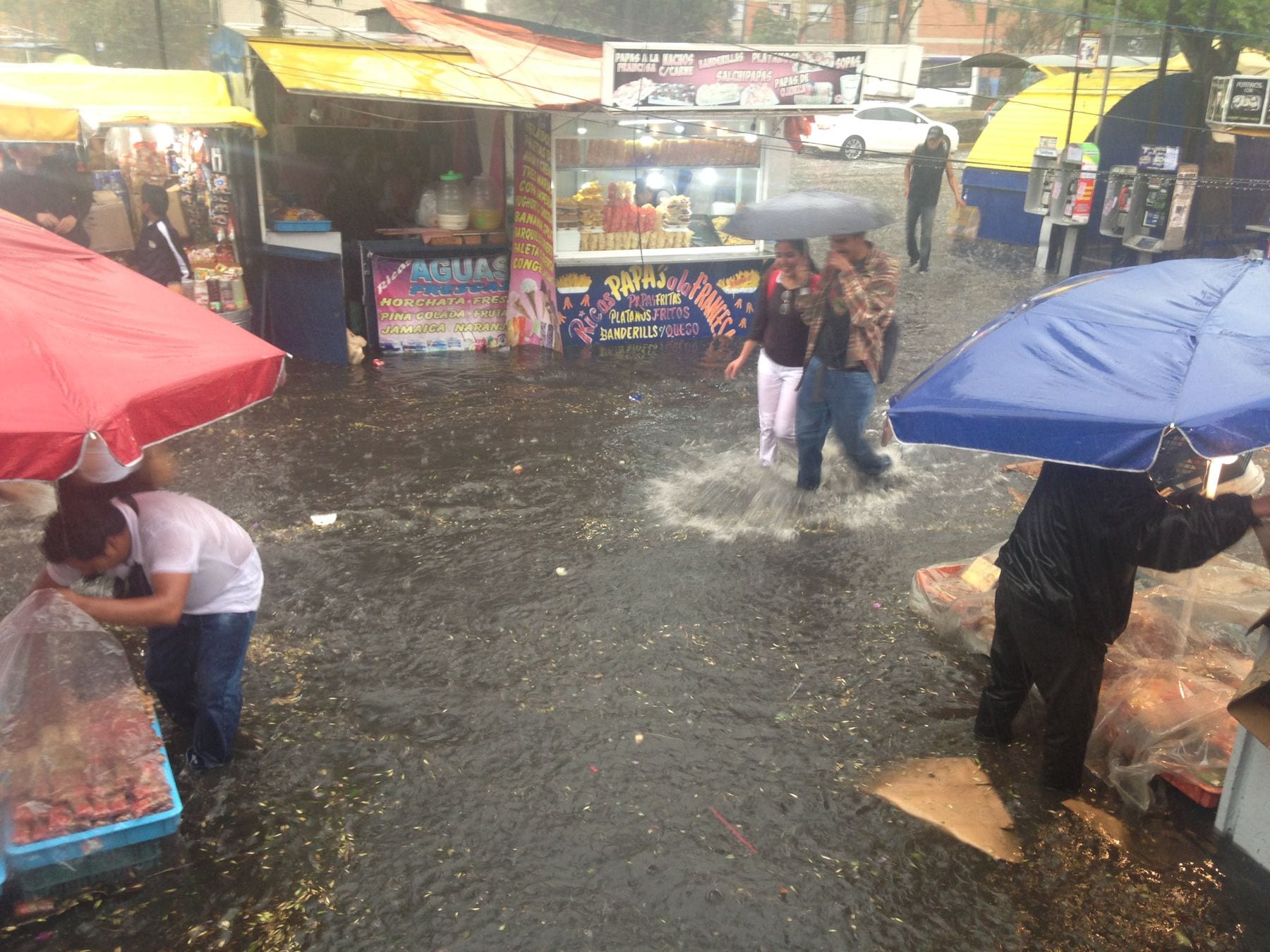 Vendedores y transeúntes cruzan una calle inundada junto a puestos de un mercado ambulante en la Ciudad de México, varios de ellos con paraguas de colores durante un aguacero.