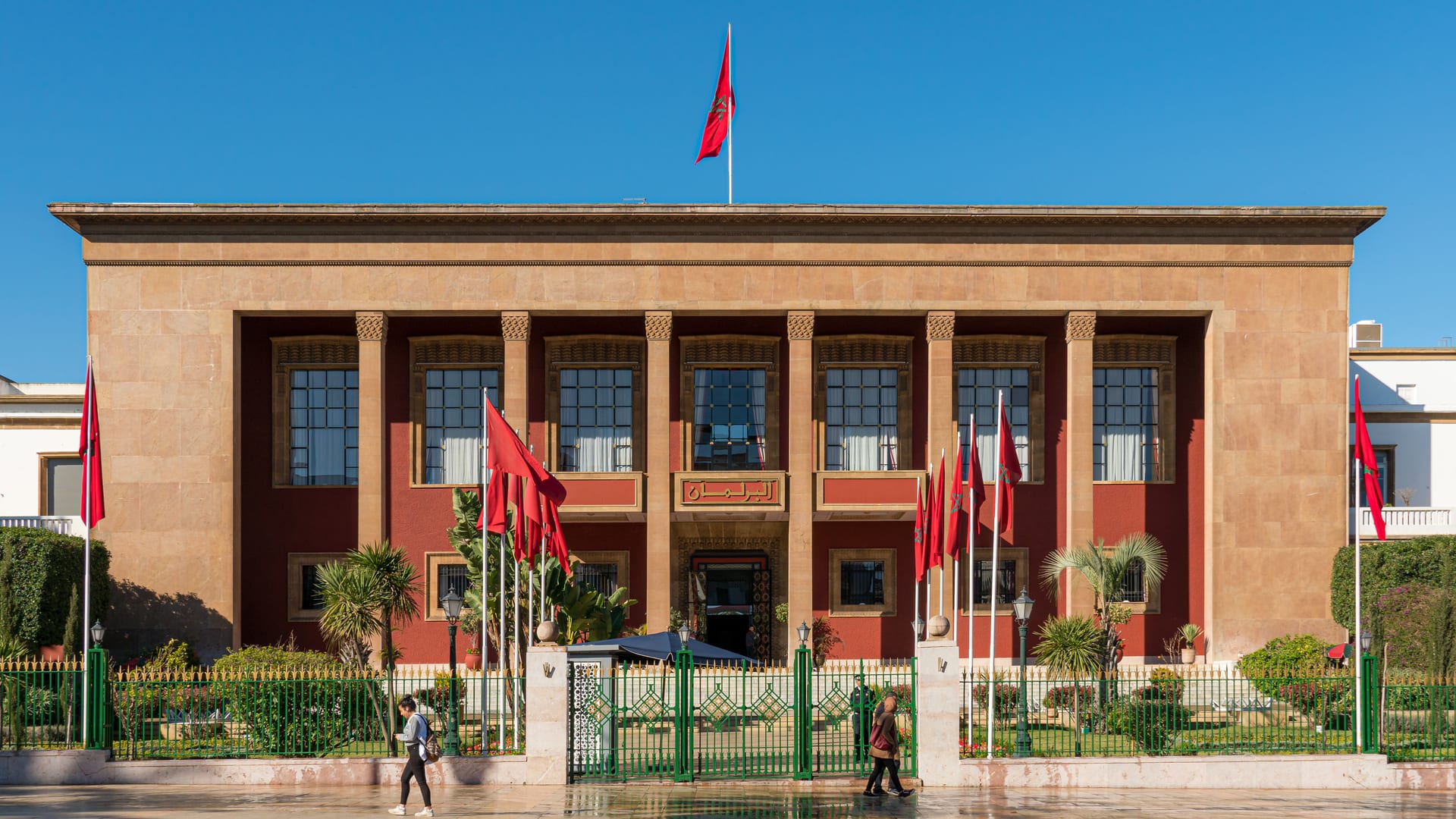 Fachada del Parlamento de Marruecos, con columnas y banderas rojas ondeando, y dos personas caminando frente a la reja de entrada.