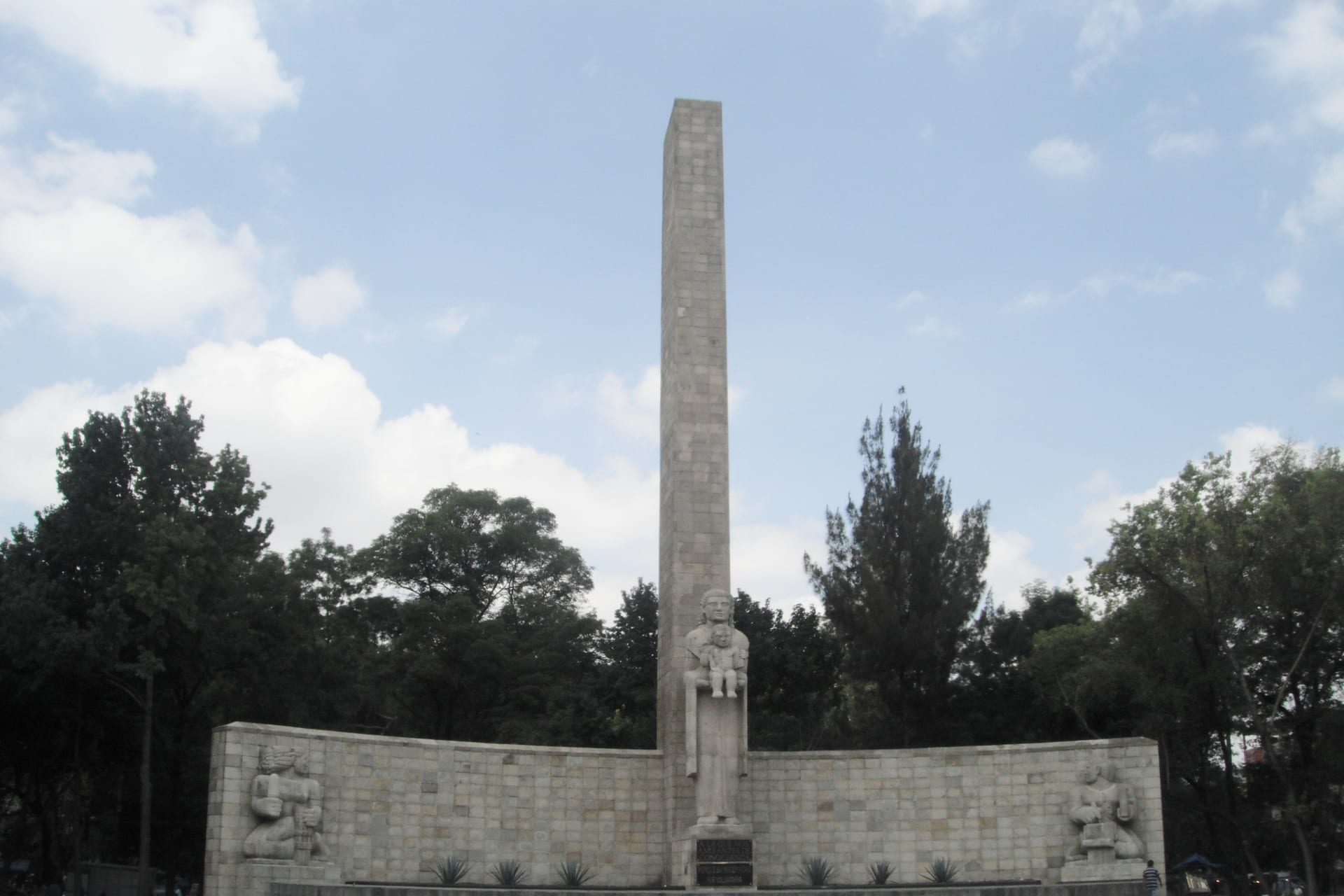El Monumento a la Madre, un obelisco de piedra con esculturas en su base, rodeado de árboles bajo un cielo con nubes.