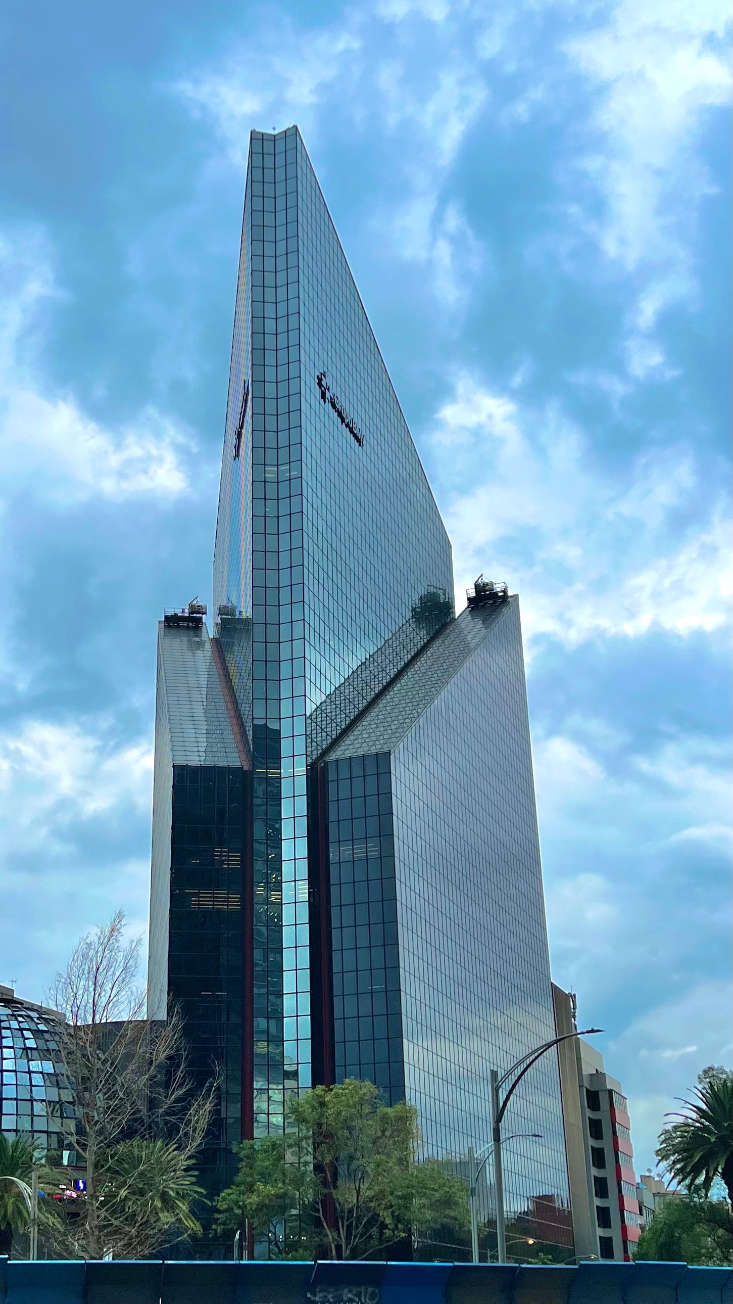 Rascacielos triangular de cristal con una cúpula curva de vidrio en su base, bajo un cielo nublado en una avenida con árboles.