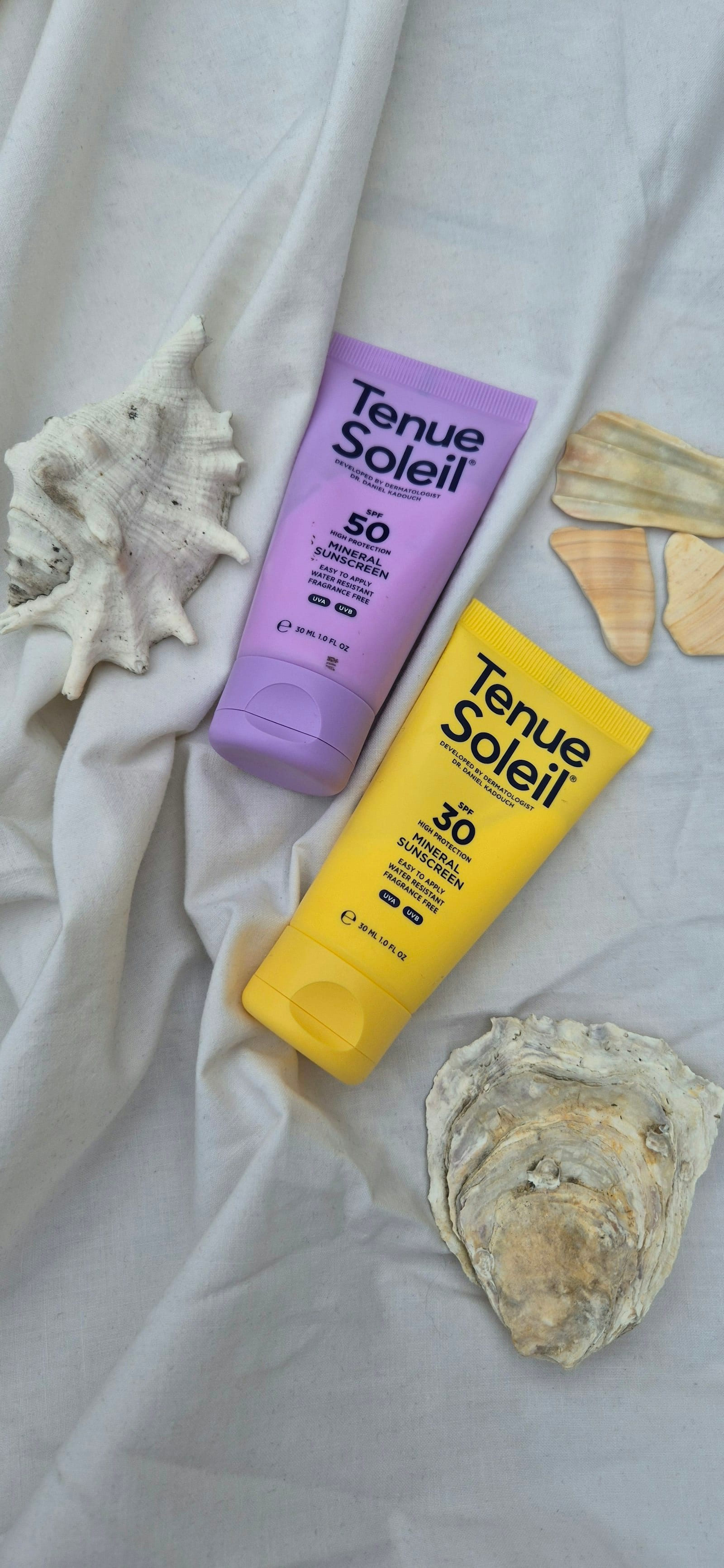 Dos tubos de protector solar mineral, uno con FPS 50 y otro con FPS 30, sobre tela blanca junto a conchas de mar.