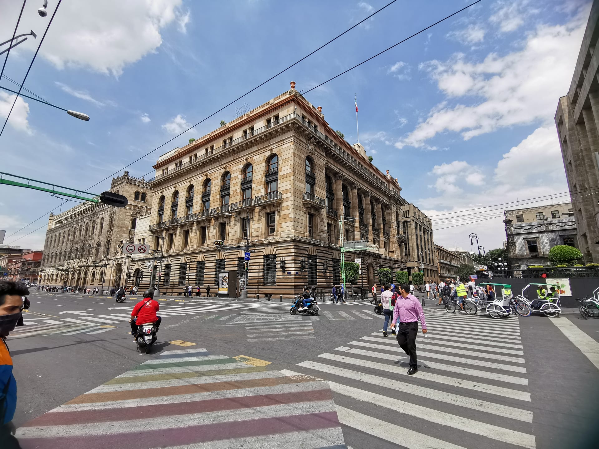 Fachada de piedra de un edificio con columnas, balcones de herrería y una bandera mexicana en la azotea, en una esquina del Centro Histórico con peatones y motociclistas cruzando.