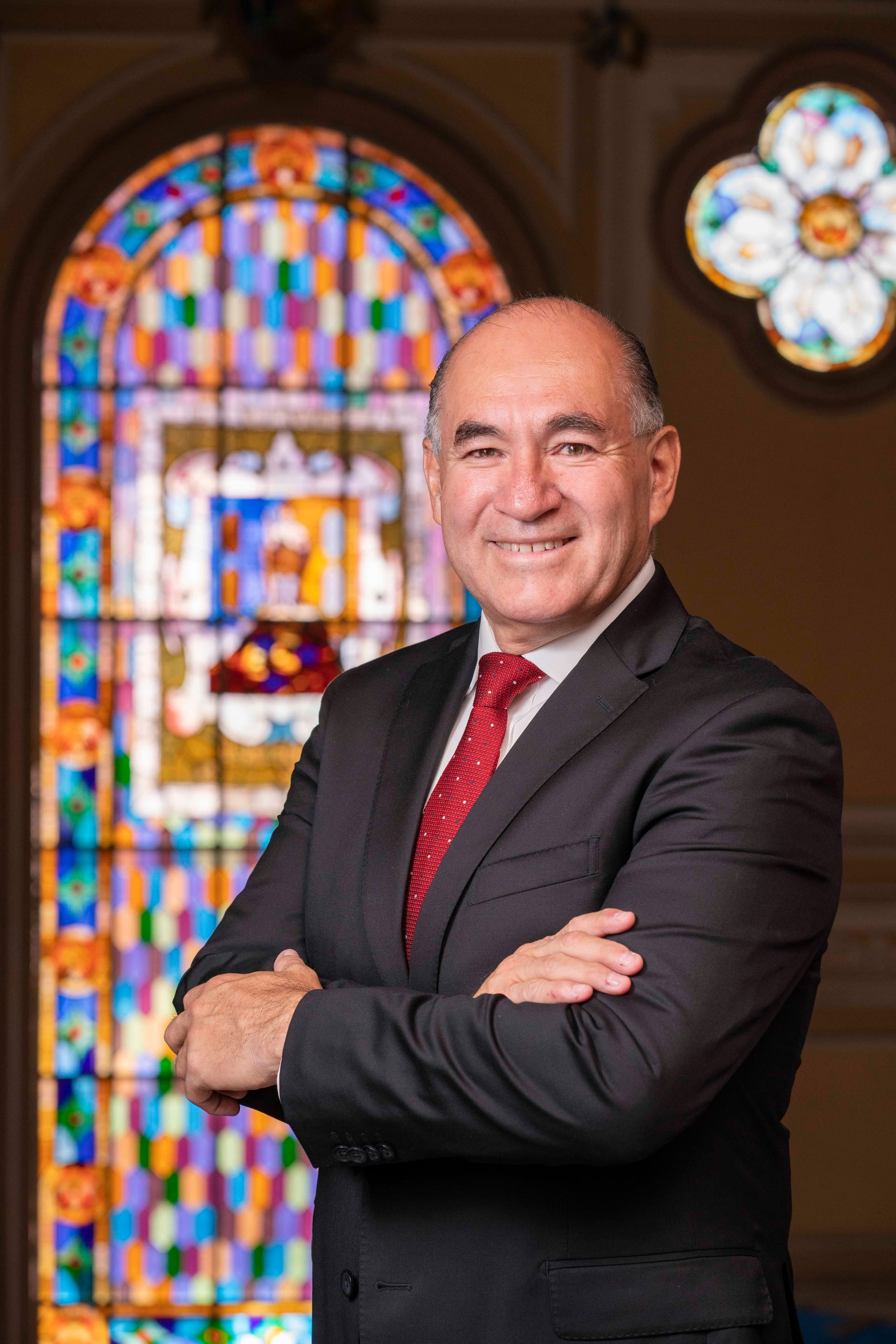 Enrique Galindo Ceballos, de traje oscuro y corbata roja, posa con los brazos cruzados frente a un vitral de colores.
