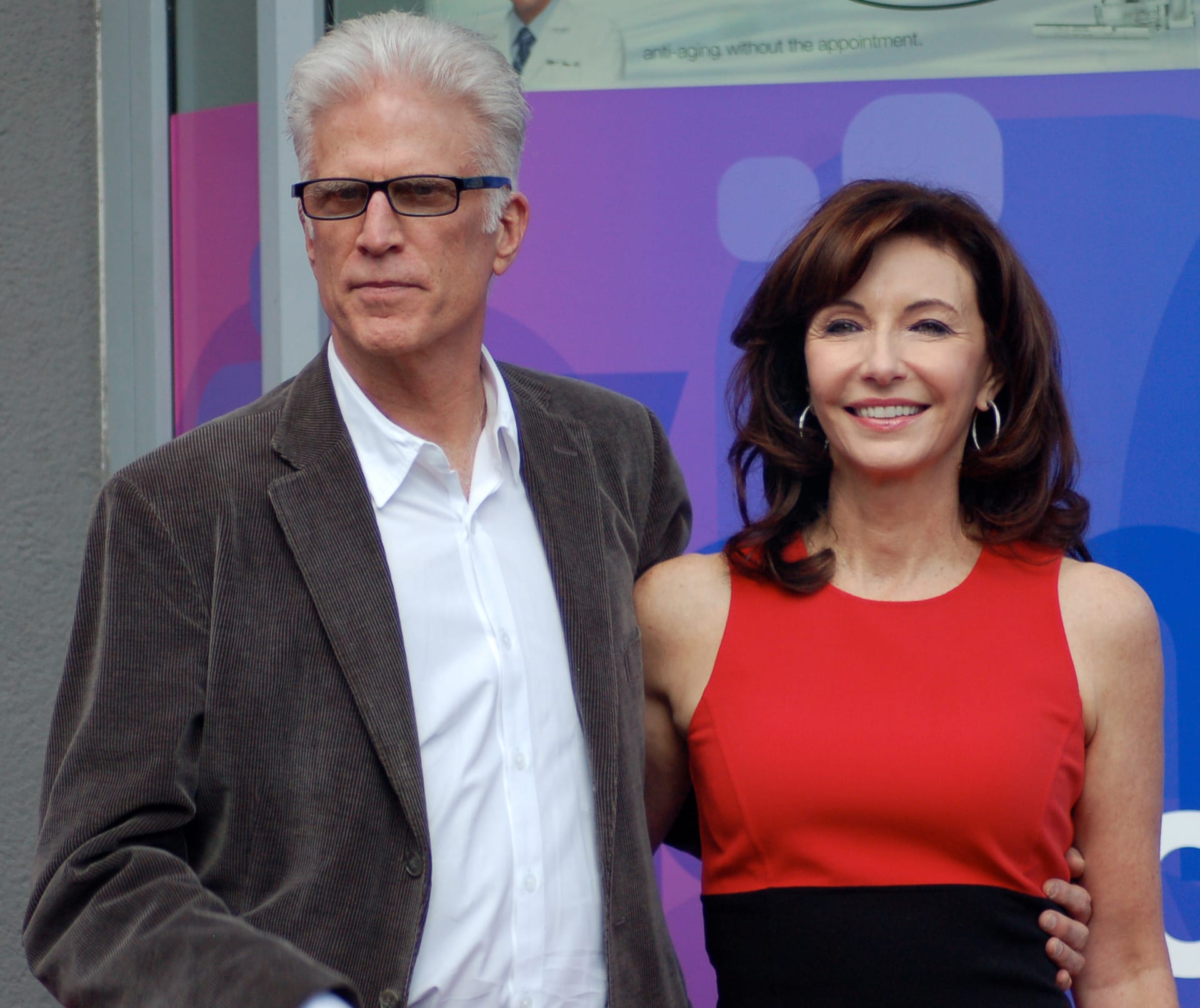 Ted Danson, con lentes y saco café, posa junto a la actriz Mary Steenburgen, quien viste un vestido rojo y negro.