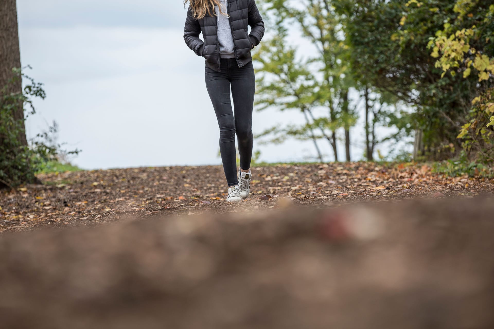 Una mujer camina por un sendero de bosque cubierto de hojas de otoño, con luz suave entre los árboles.