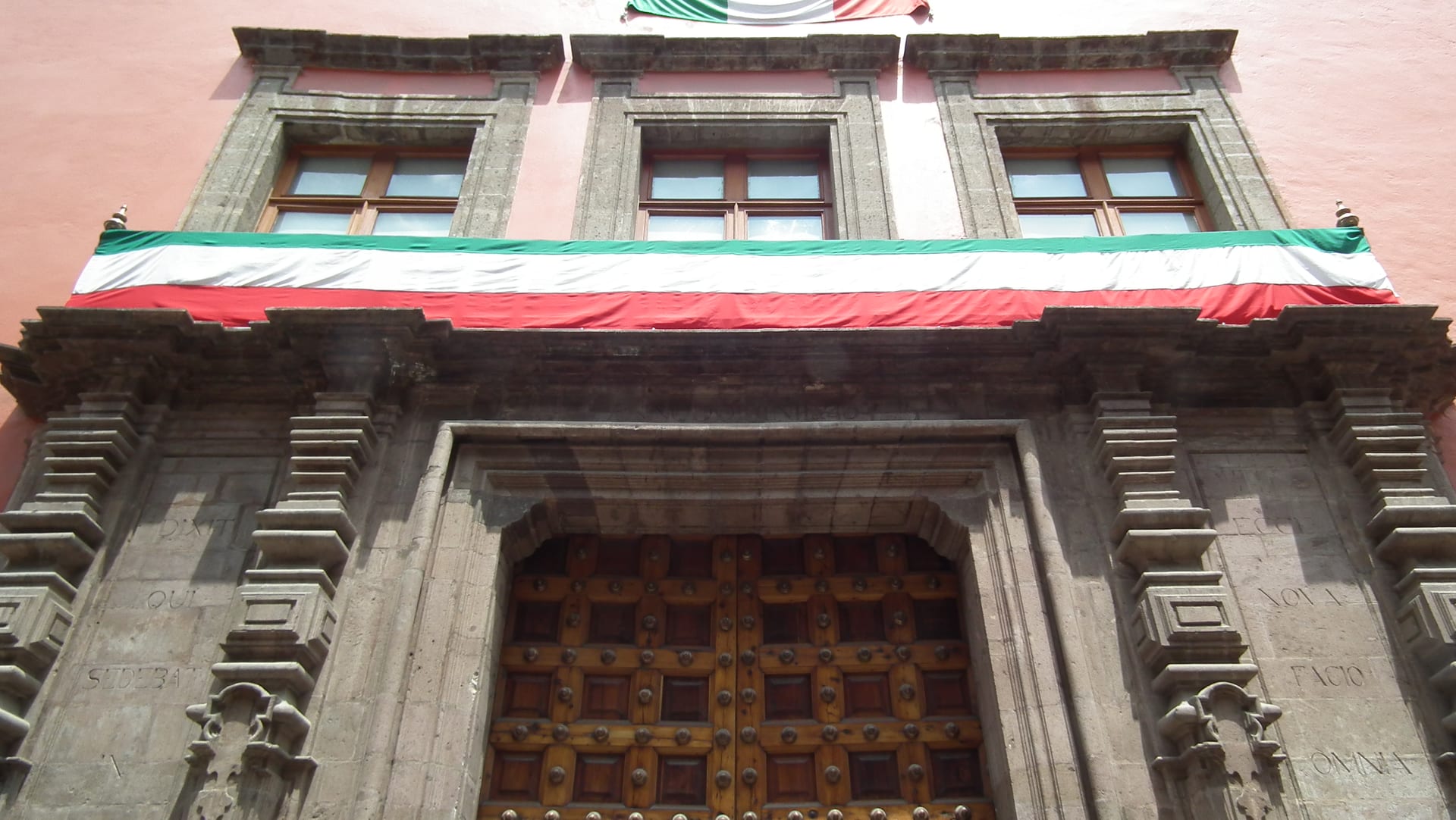 Fachada de cantera del antiguo Palacio del Arzobispado, sede del Museo de la SHCP, con una bandera mexicana colgada sobre el balcón y un portón de madera con tachuelas.