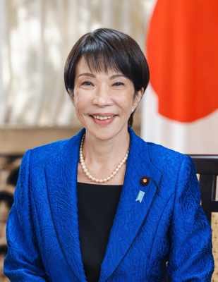Retrato oficial de una mujer de cabello corto oscuro y traje azul, de pie frente a la bandera de Japón.