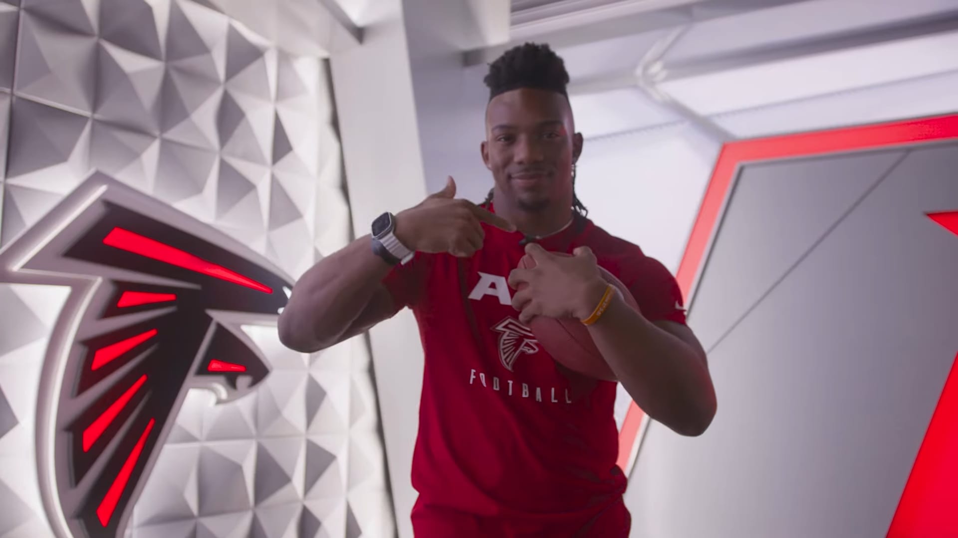 Bijan Robinson sonríe y señala su brazalete mientras sostiene un balón, frente a un muro con el logotipo de los Falcons.