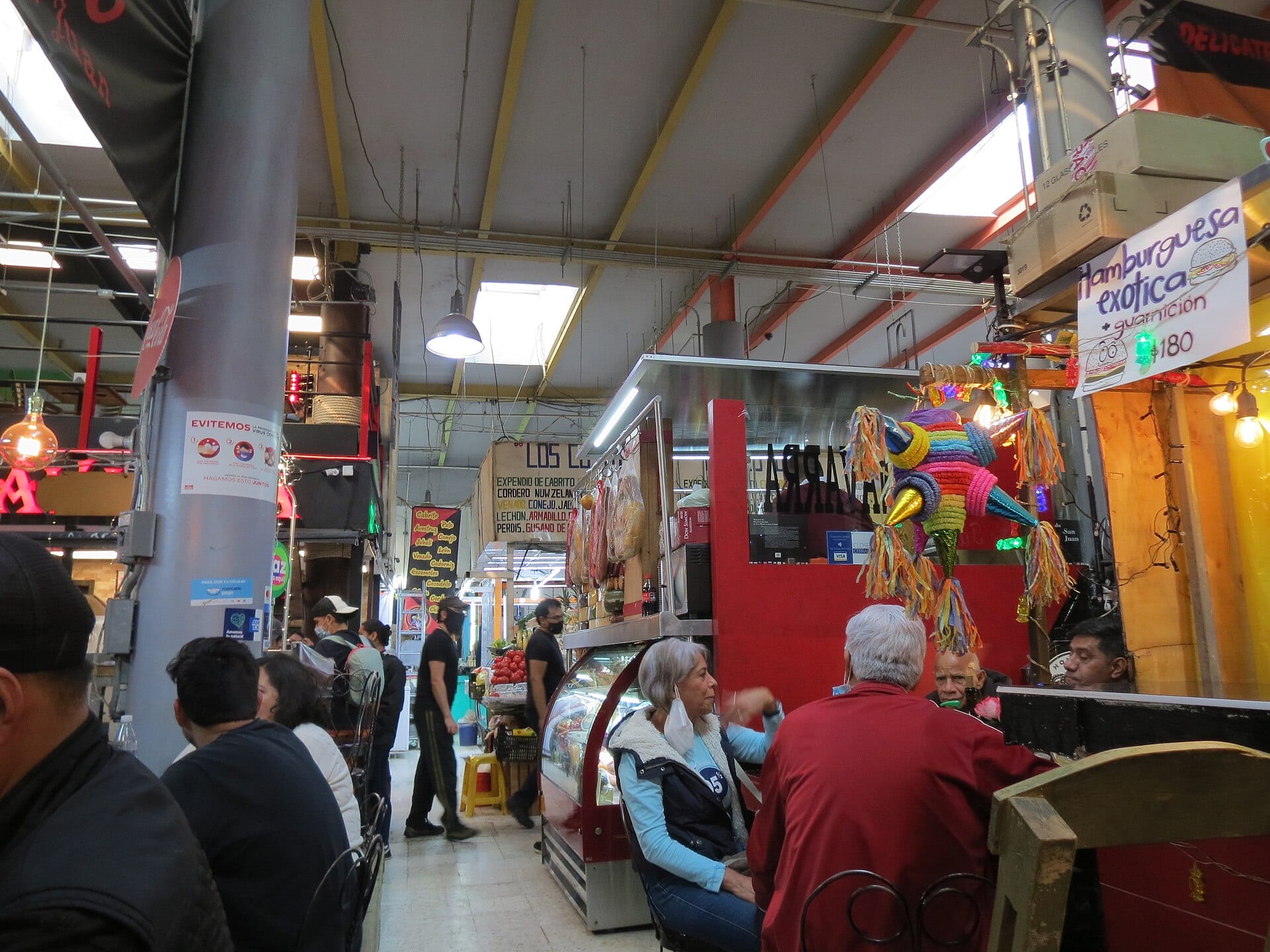 Comensales sentados junto a los puestos de un mercado techado, con vitrinas refrigeradas y una piñata colgada sobre uno de los locales.