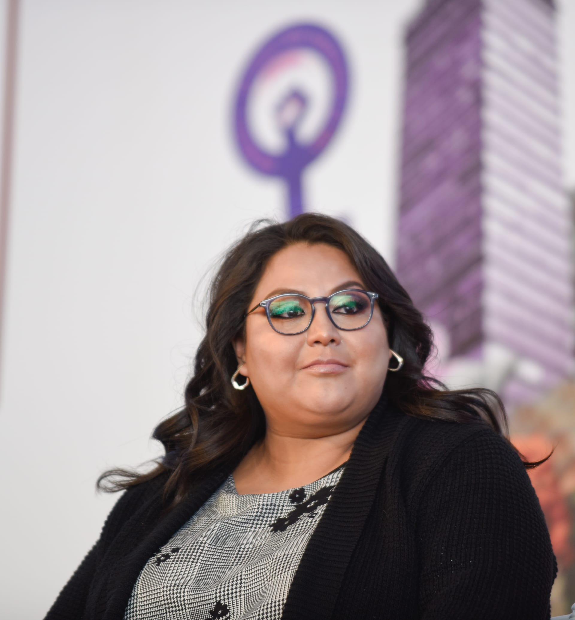 Mujer con lentes y cabello ondulado oscuro, con arracadas y suéter negro, frente al símbolo morado del movimiento feminista.