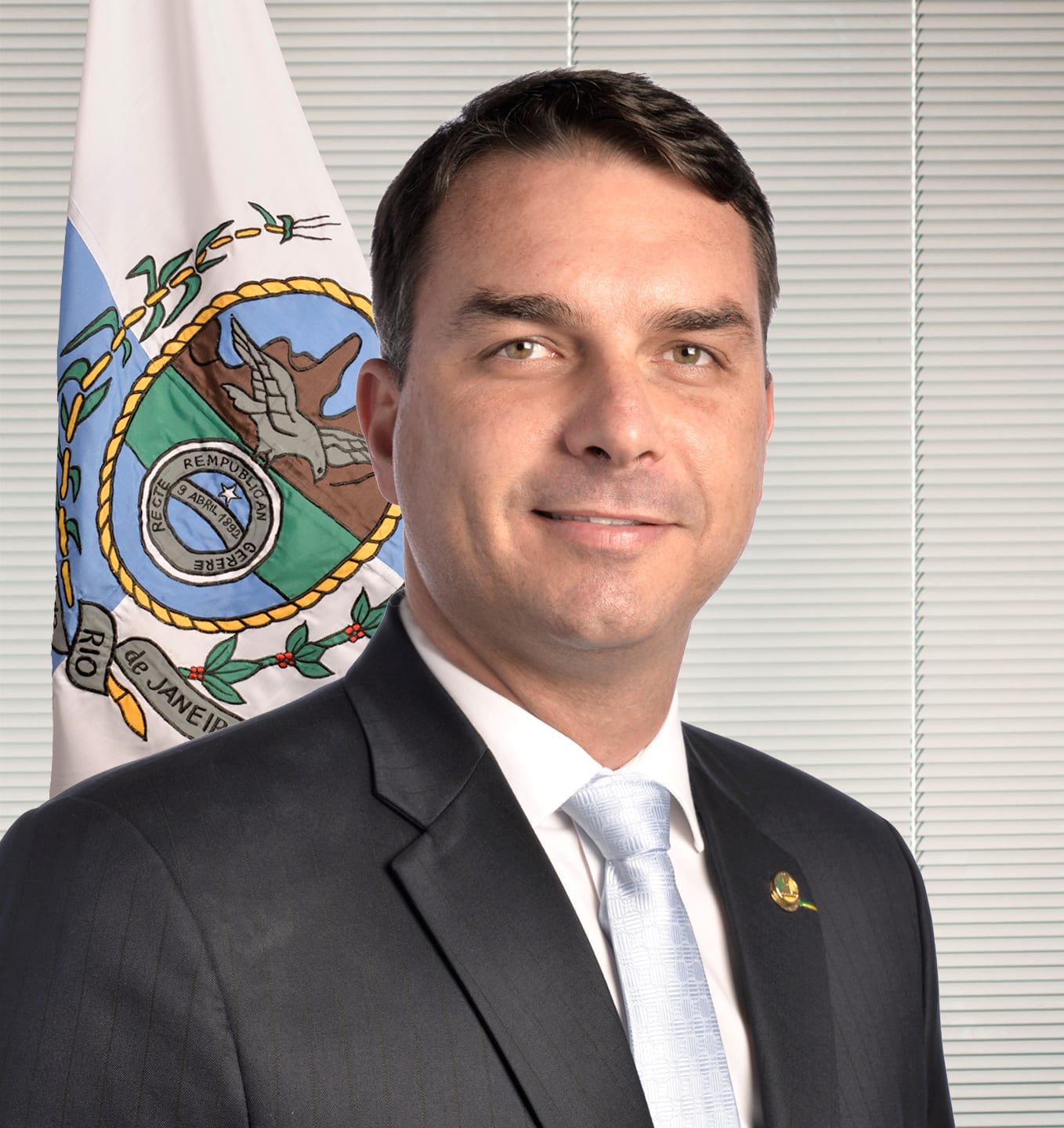 Flávio Bolsonaro, de traje oscuro y corbata azul claro, frente a la bandera de Río de Janeiro.