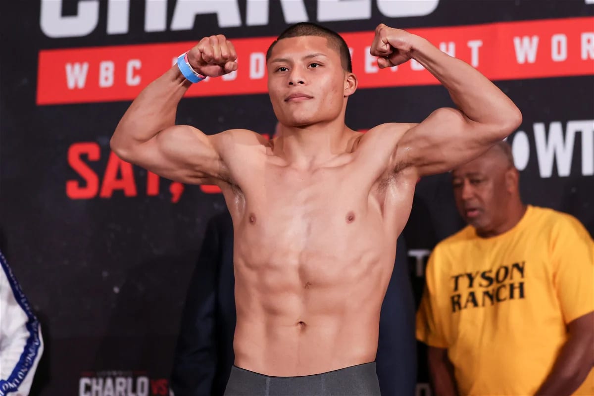 Isaac "Pitbull" Cruz, sin camisa, flexiona los brazos durante una ceremonia de pesaje anterior, con un cartel del Consejo Mundial de Boxeo al fondo.