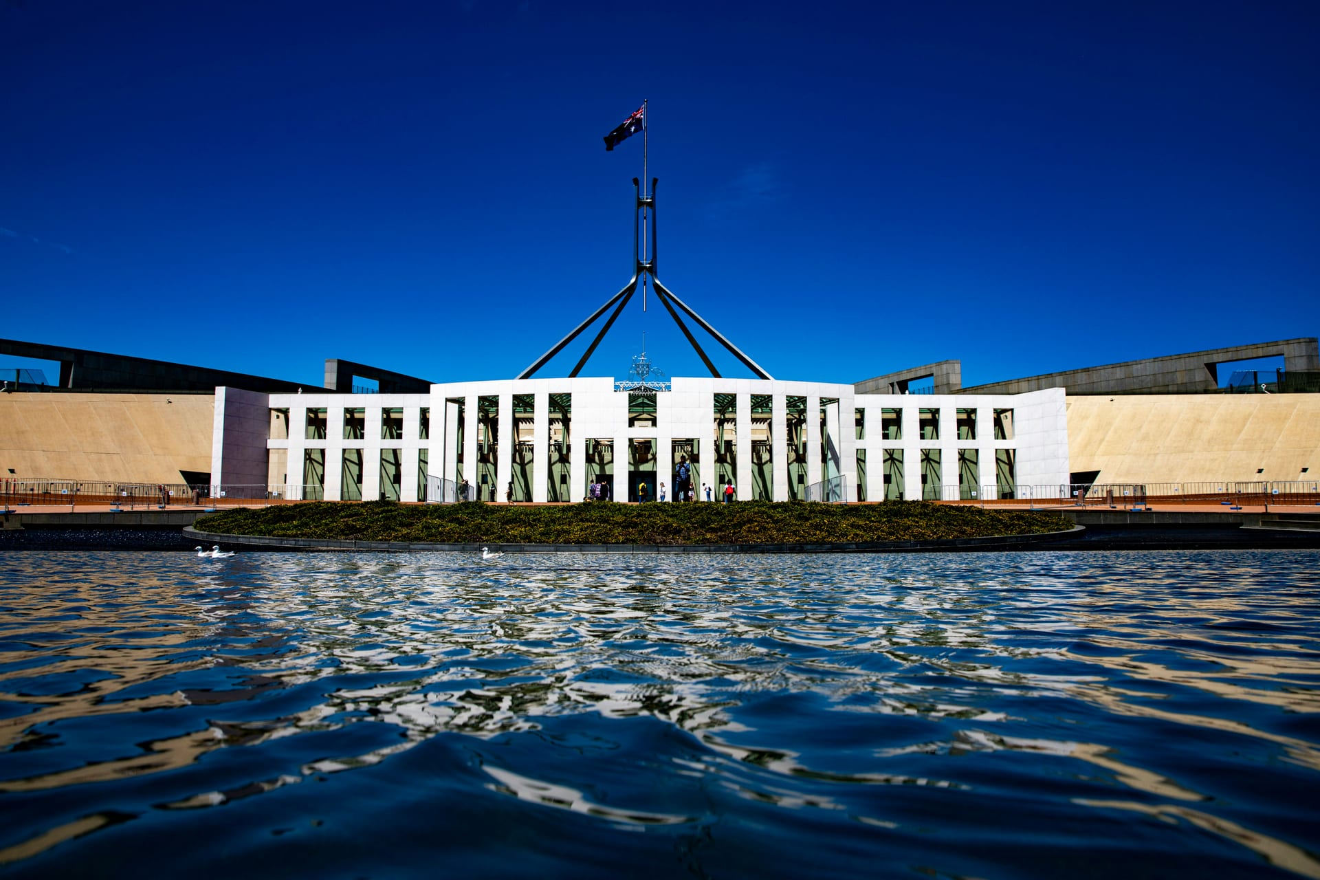 El edificio del Parlamento de Australia en Canberra, con su asta bandera y un espejo de agua al frente.
