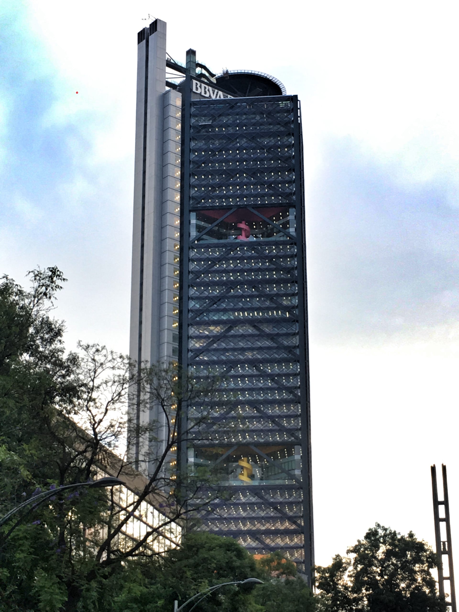 Torre de vidrio azulado con el logotipo de BBVA en lo alto, fotografiada desde la calle entre árboles.