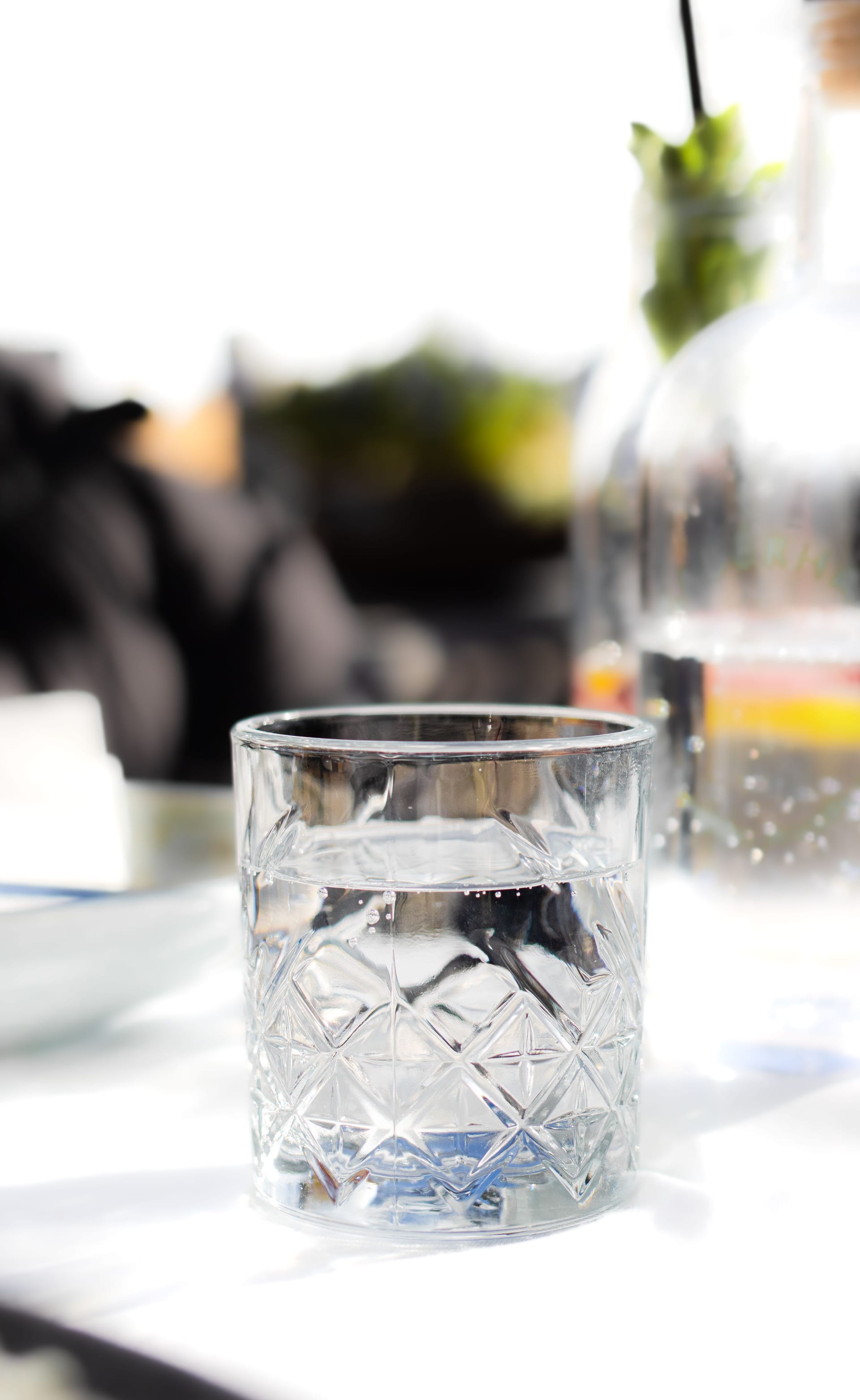 Un vaso de cristal tallado, lleno hasta la mitad de agua, sobre una mesa blanca con objetos desenfocados al fondo.