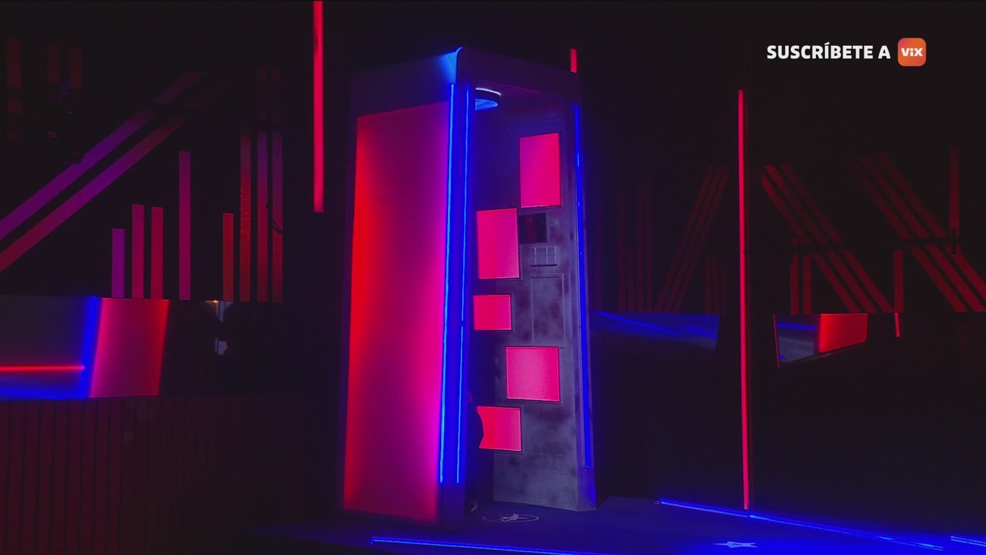 Una estructura tipo cabina con paneles rosas y bordes de neón azul, dentro de un cuarto oscuro con líneas de luz roja y azul.