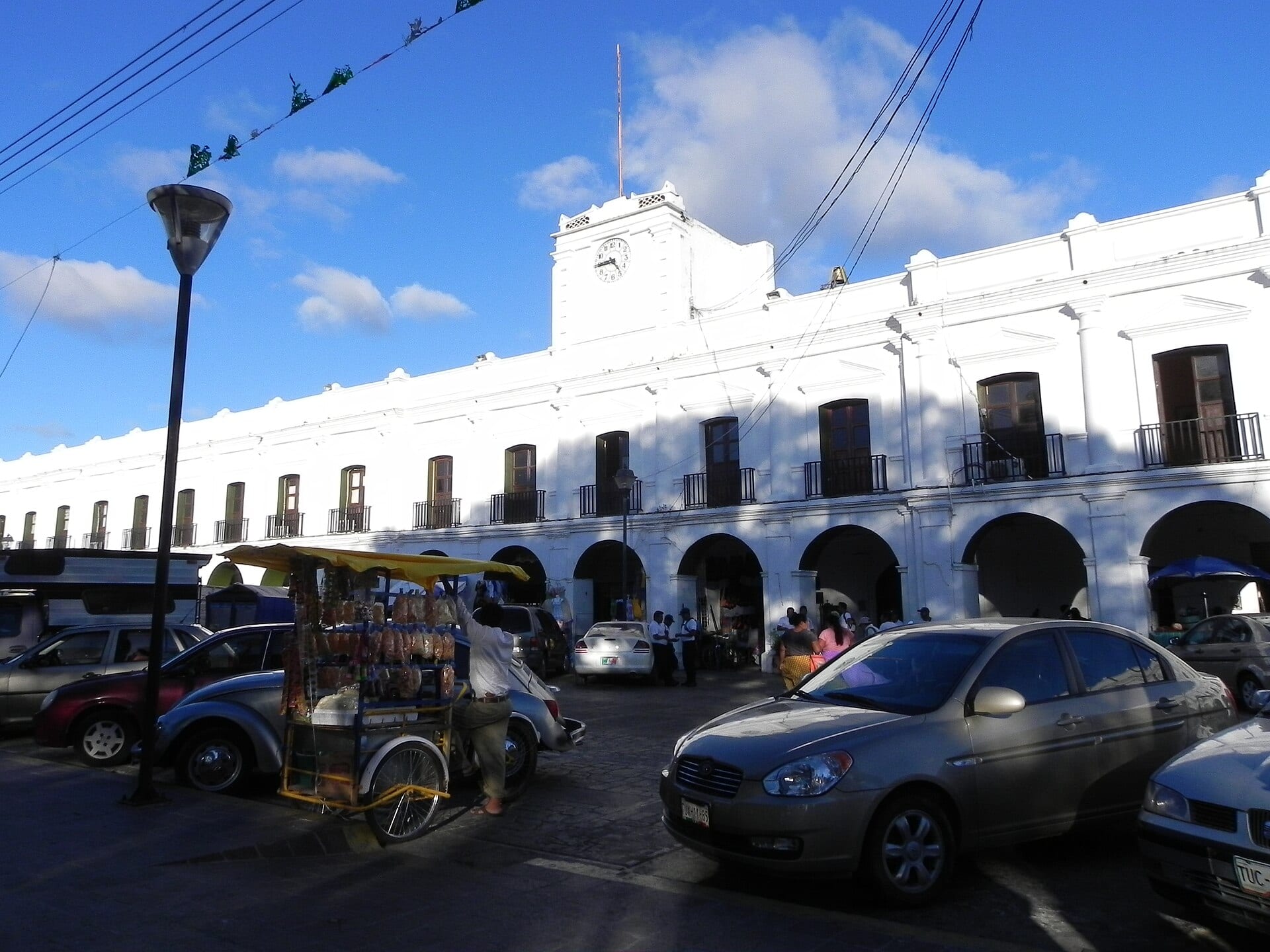 El Palacio Municipal de Juchitán de Zaragoza, un edificio colonial blanco con arcadas y un reloj en la torre, con autos estacionados al frente.