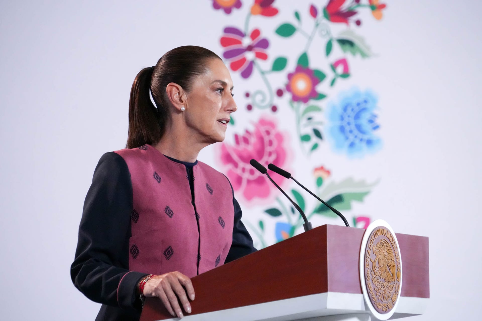 Claudia Sheinbaum habla de perfil frente a dos micrófonos, con un fondo floral bordado detrás.