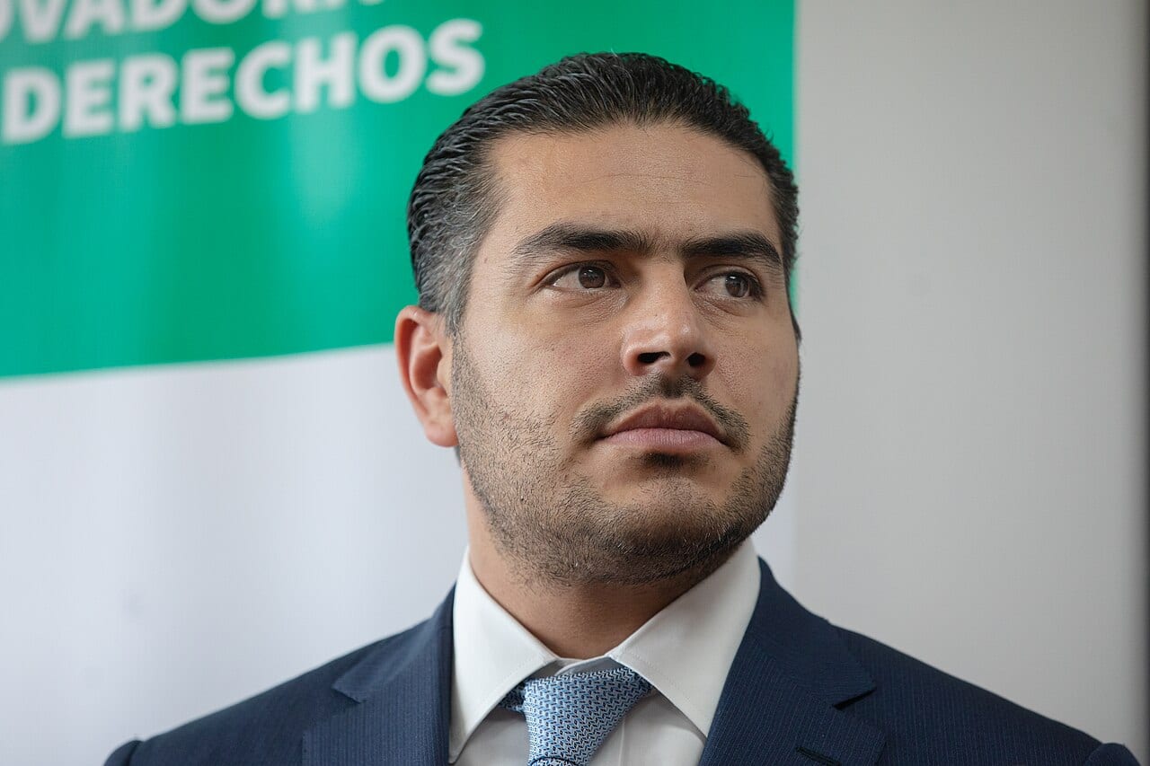 Omar García Harfuch, con traje azul marino y corbata clara a rayas, mira hacia un lado frente a un cartel verde y blanco con la palabra Derechos visible detrás.