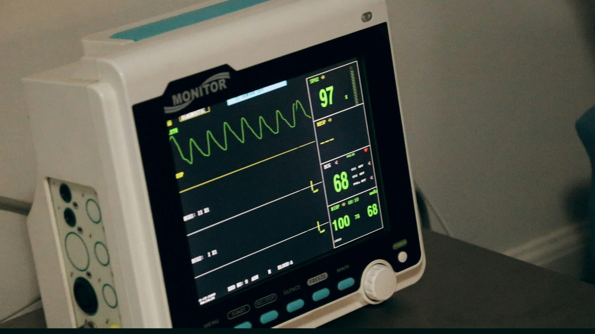 Monitor médico verde muestra trazos ondulantes de un electrocardiograma en un cuarto en penumbra.