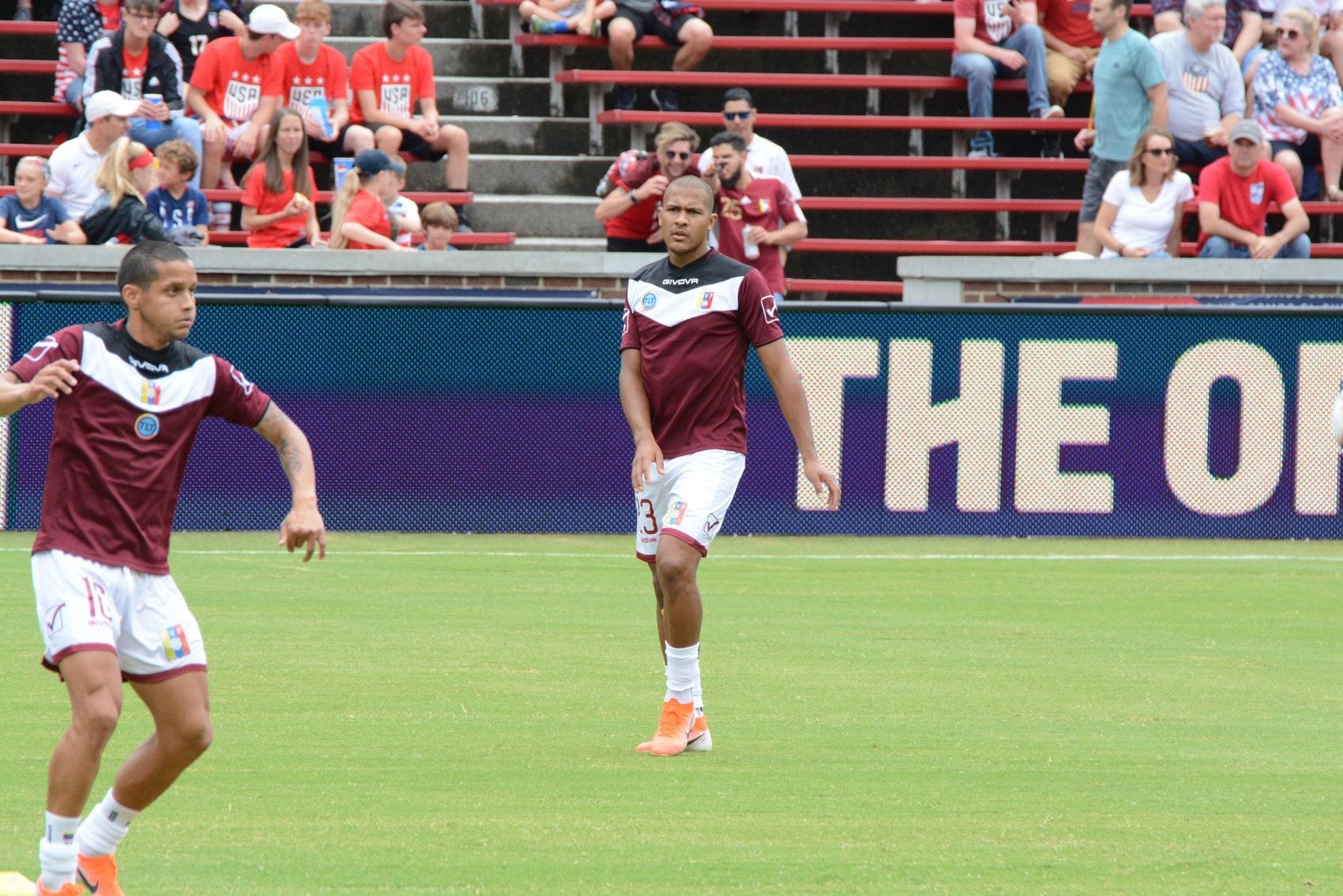 Un futbolista con uniforme vinotinto de Venezuela camina en la cancha durante un calentamiento, con un compañero a un costado y aficionados en las gradas al fondo, en una imagen de archivo con su selección.