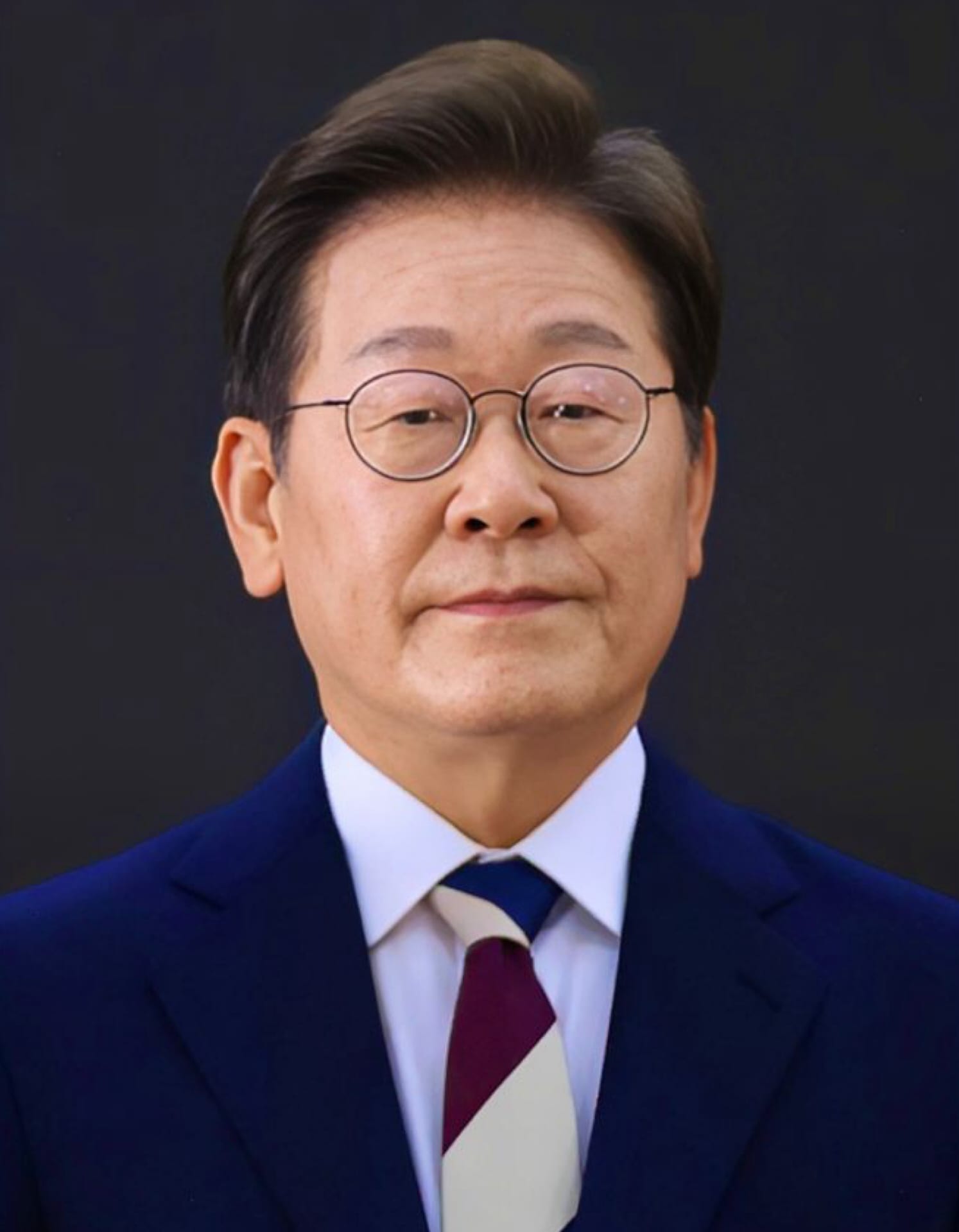 Lee Jae-myung, con traje oscuro y corbata a rayas azul y vino, posa de frente ante un fondo oscuro.