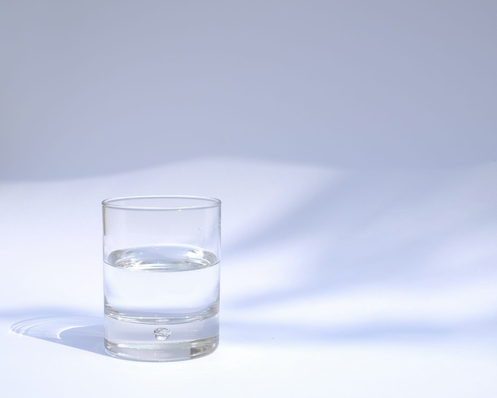 Un vaso de vidrio transparente lleno de agua hasta la mitad, sobre una superficie blanca con luz natural.