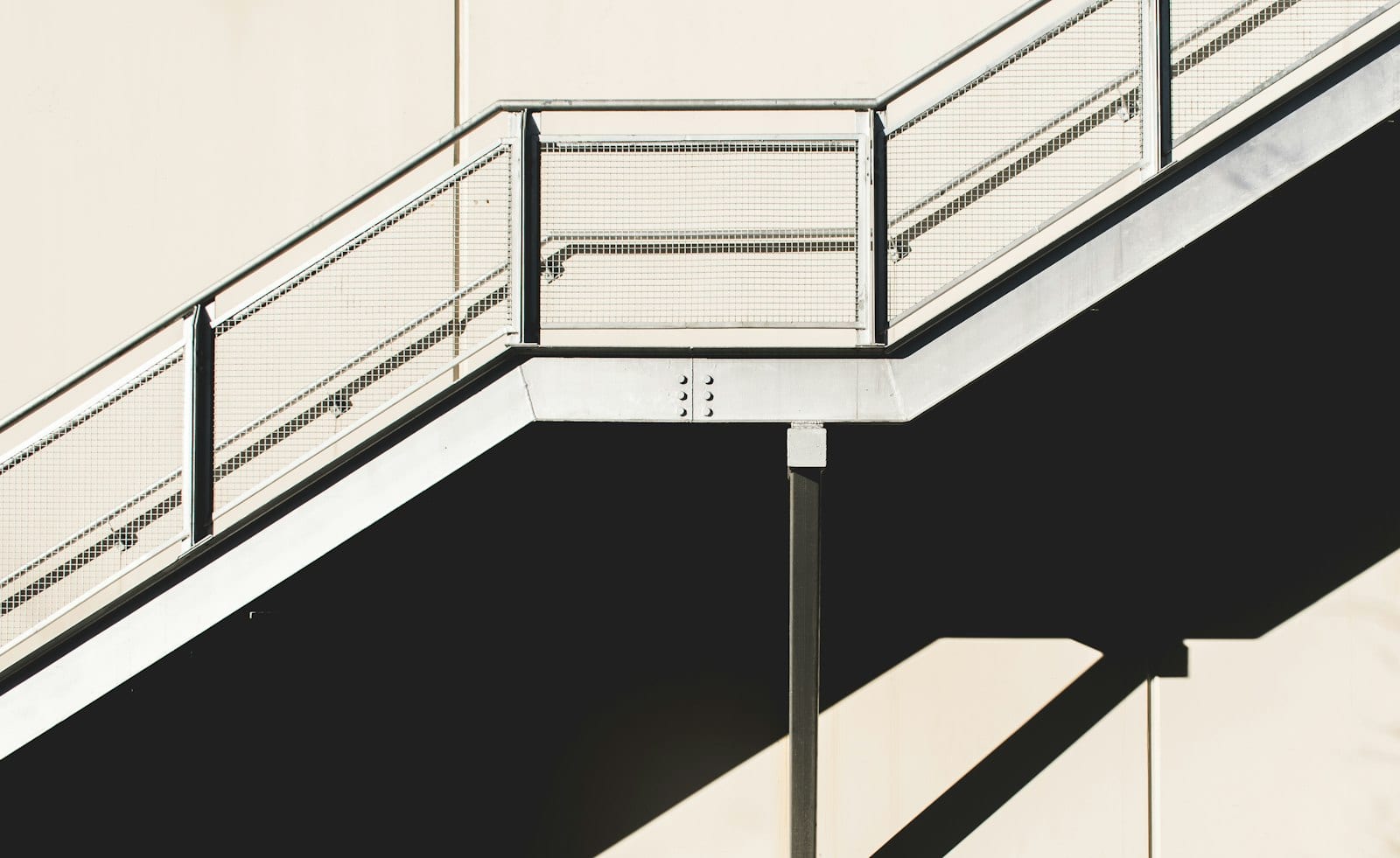 Escalera metálica exterior con barandal de malla, vista desde abajo con sombras marcadas.