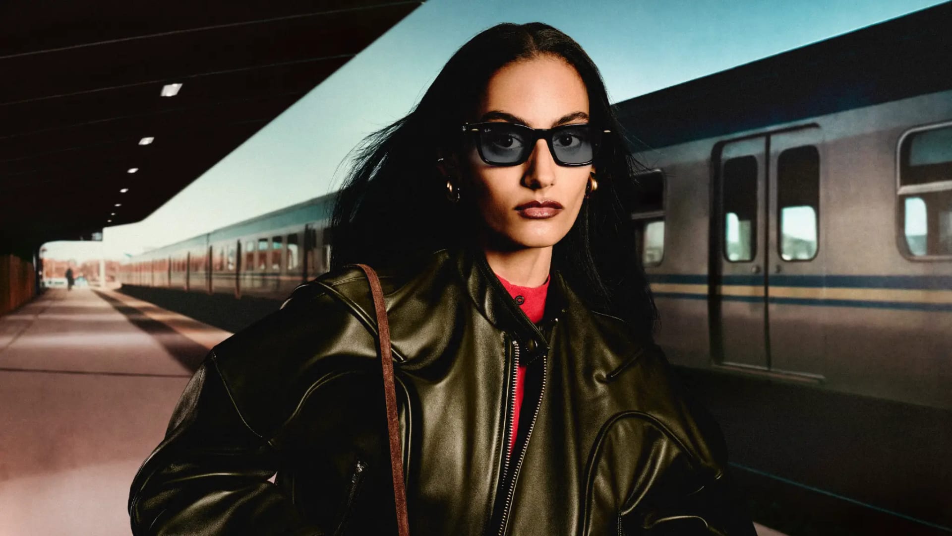 Una mujer con gafas de sol negras de armazón grueso y chamarra de piel espera en un andén de tren.