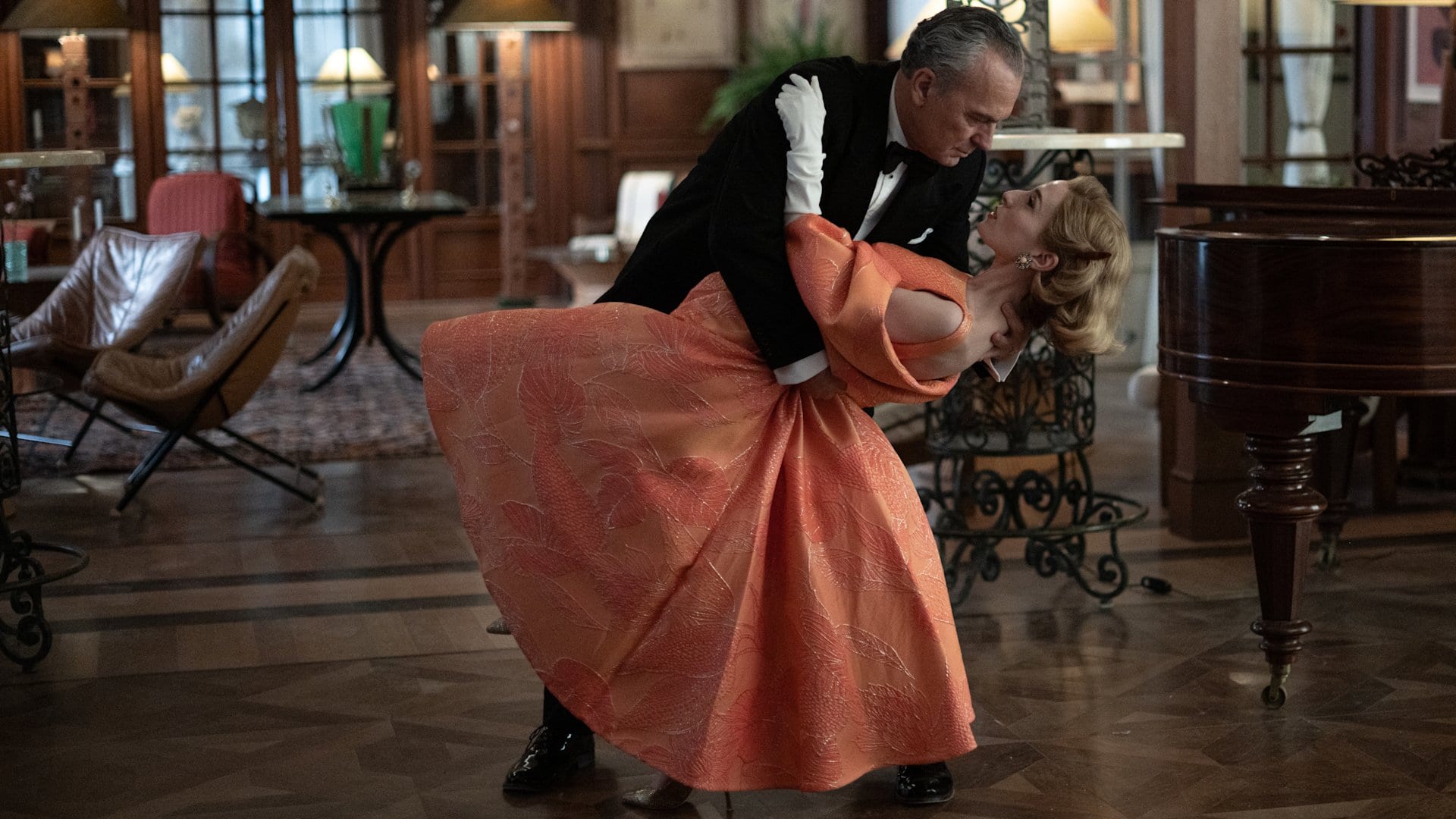 Un actor de esmoquin negro dobla hacia atrás a una actriz de vestido color durazno en un paso de baile, dentro de un salón con piano de cola.
