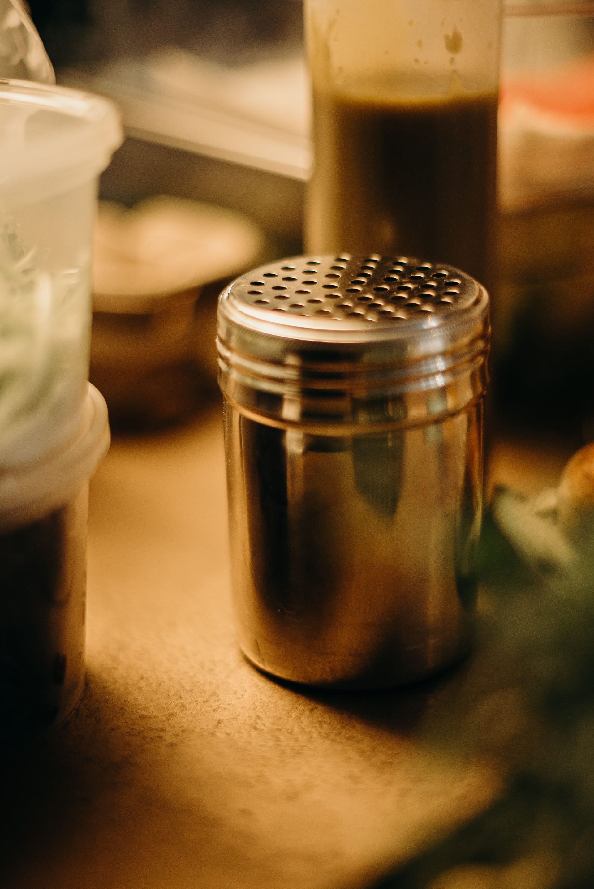 Un shaker metálico con tapa perforada sobre una mesa de madera, entre botellas de aderezo y otros utensilios de cocina.