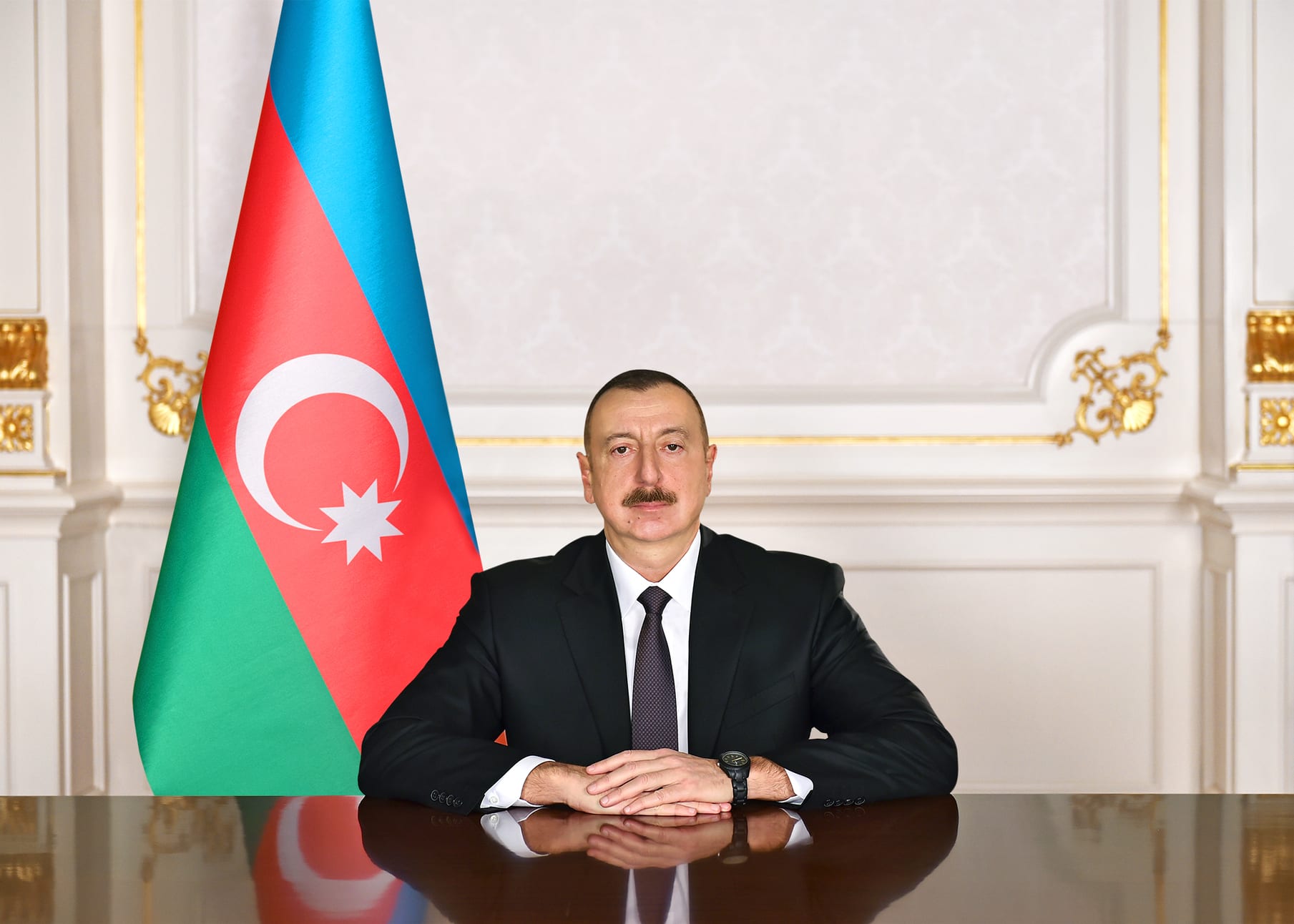 Ilham Aliyev, de traje oscuro, sentado a una mesa junto a la bandera de Azerbaiyán, con las manos entrelazadas frente a él.