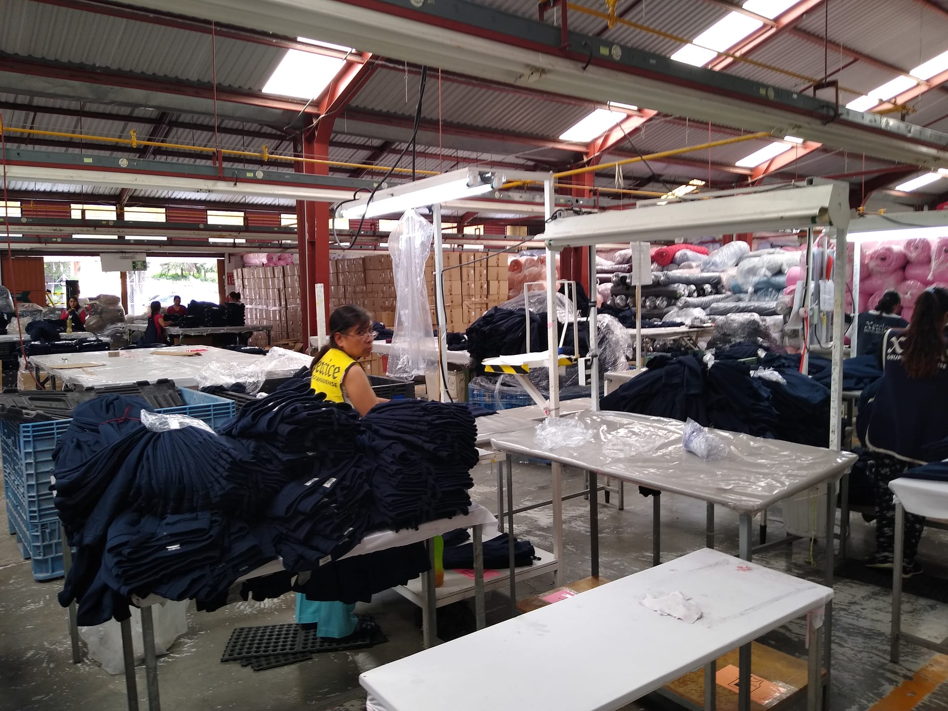Una trabajadora con chaleco amarillo dobla prendas azul marino junto a mesas cargadas de ropa doblada y rollos de tela.