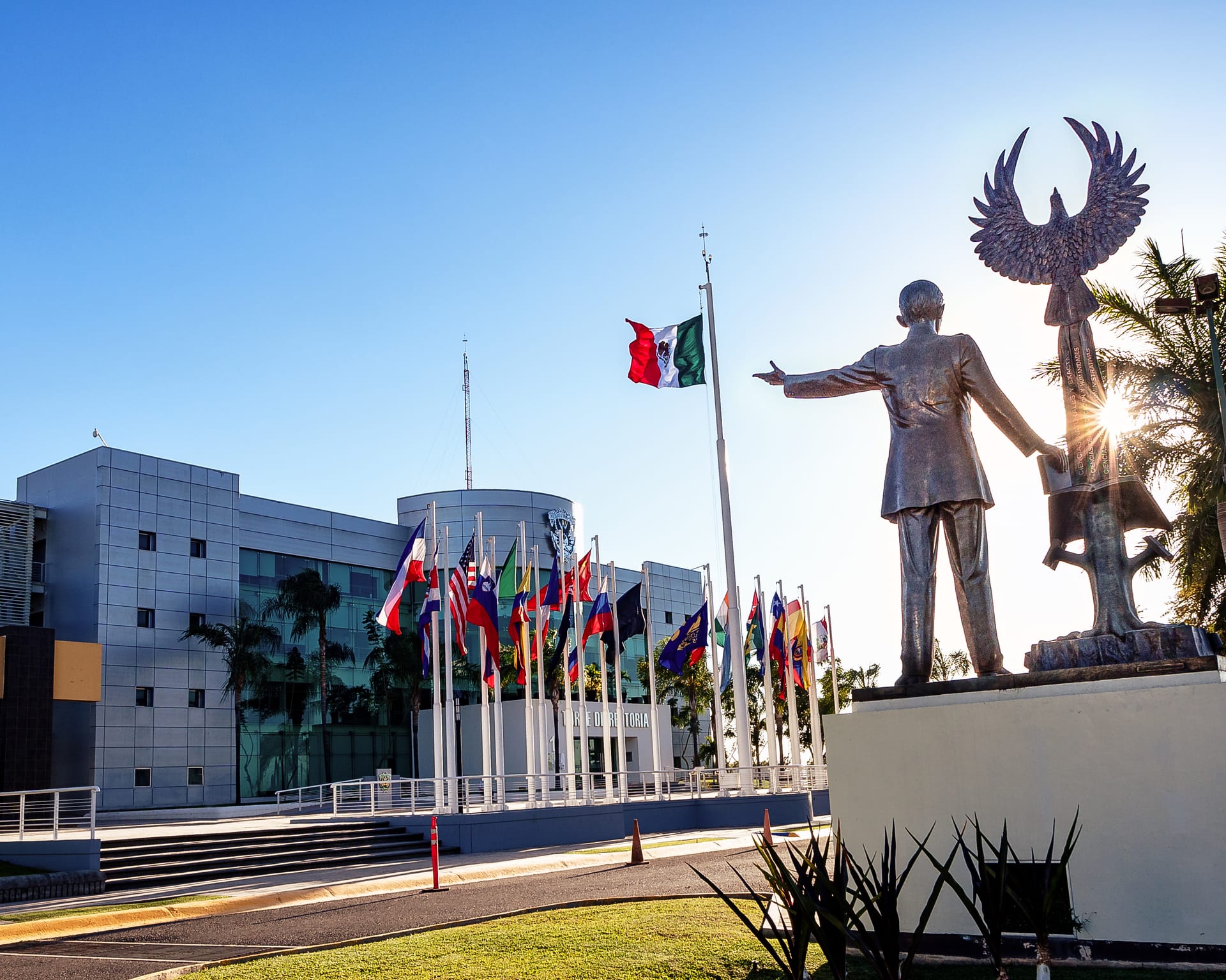 La Torre de Rectoría del campus Rafael Buelna de la Universidad Autónoma de Sinaloa, en Culiacán, con una fila de banderas y una escultura al frente.