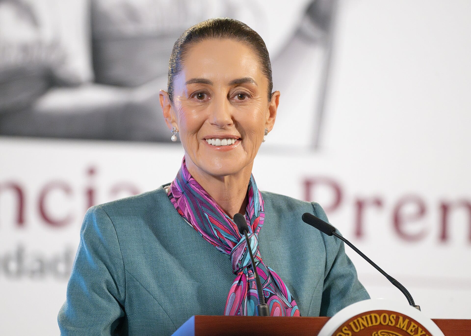 Claudia Sheinbaum sonríe detrás de un atril con el escudo de México y dos micrófonos, durante un evento oficial.