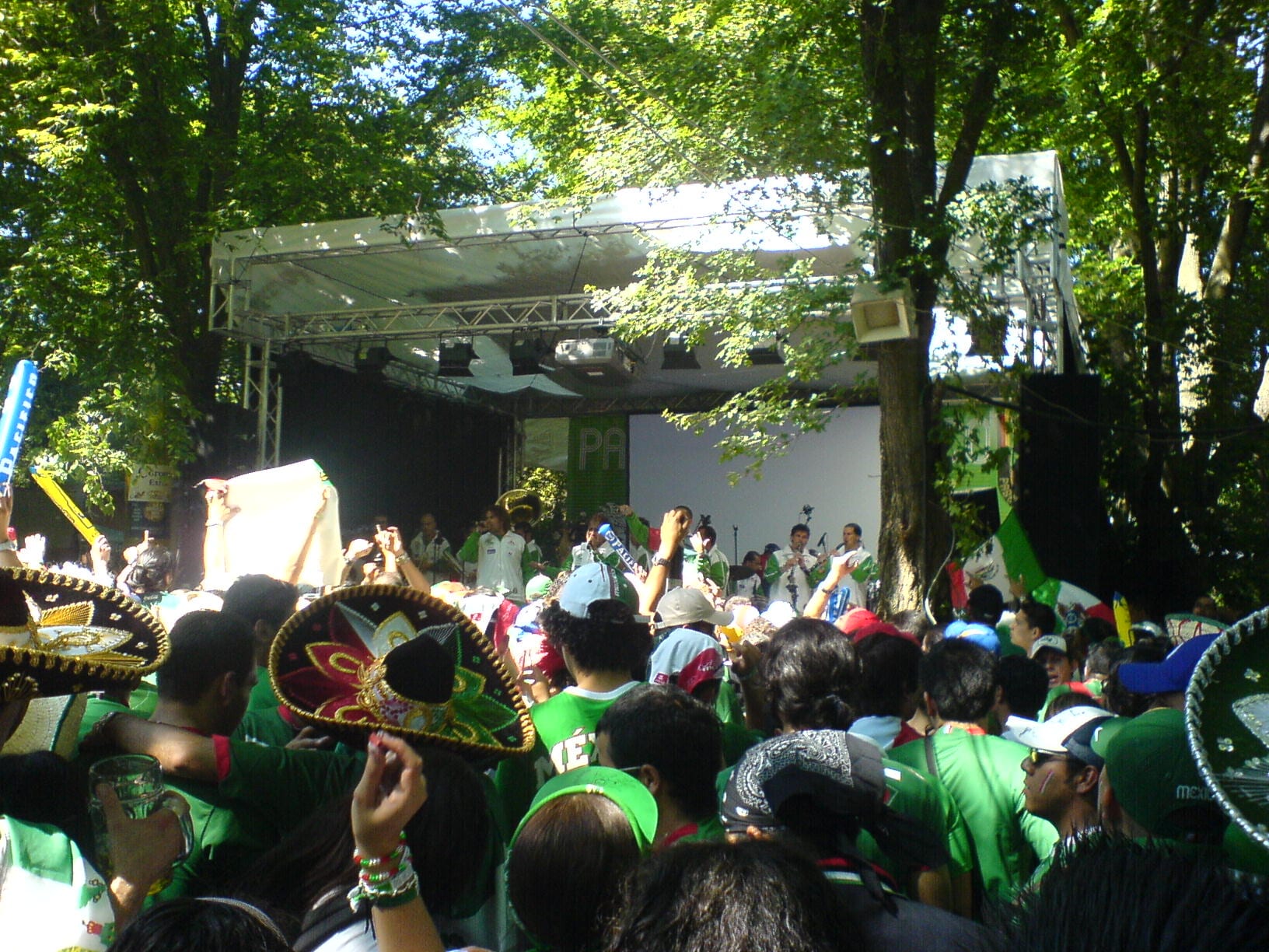 Una multitud con playeras verdes y sombreros mexicanos observa a Banda El Recodo tocar en un escenario entre los árboles, en un Fanfest del Mundial en Alemania.