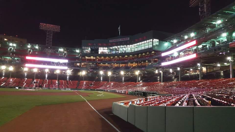 Fenway Park iluminado de noche, con las gradas vacías, el diamante de béisbol y las torres de luz encendidas.