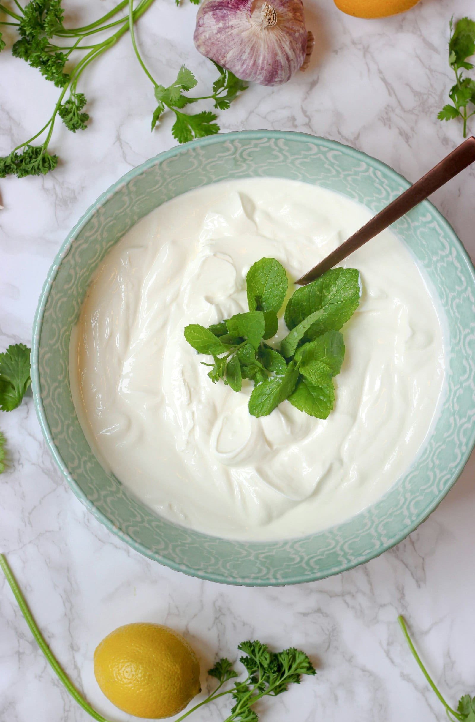 Tazón de yogur natural con hojas de menta al centro y una cuchara metálica, rodeado de ajo, limón y hierbas.